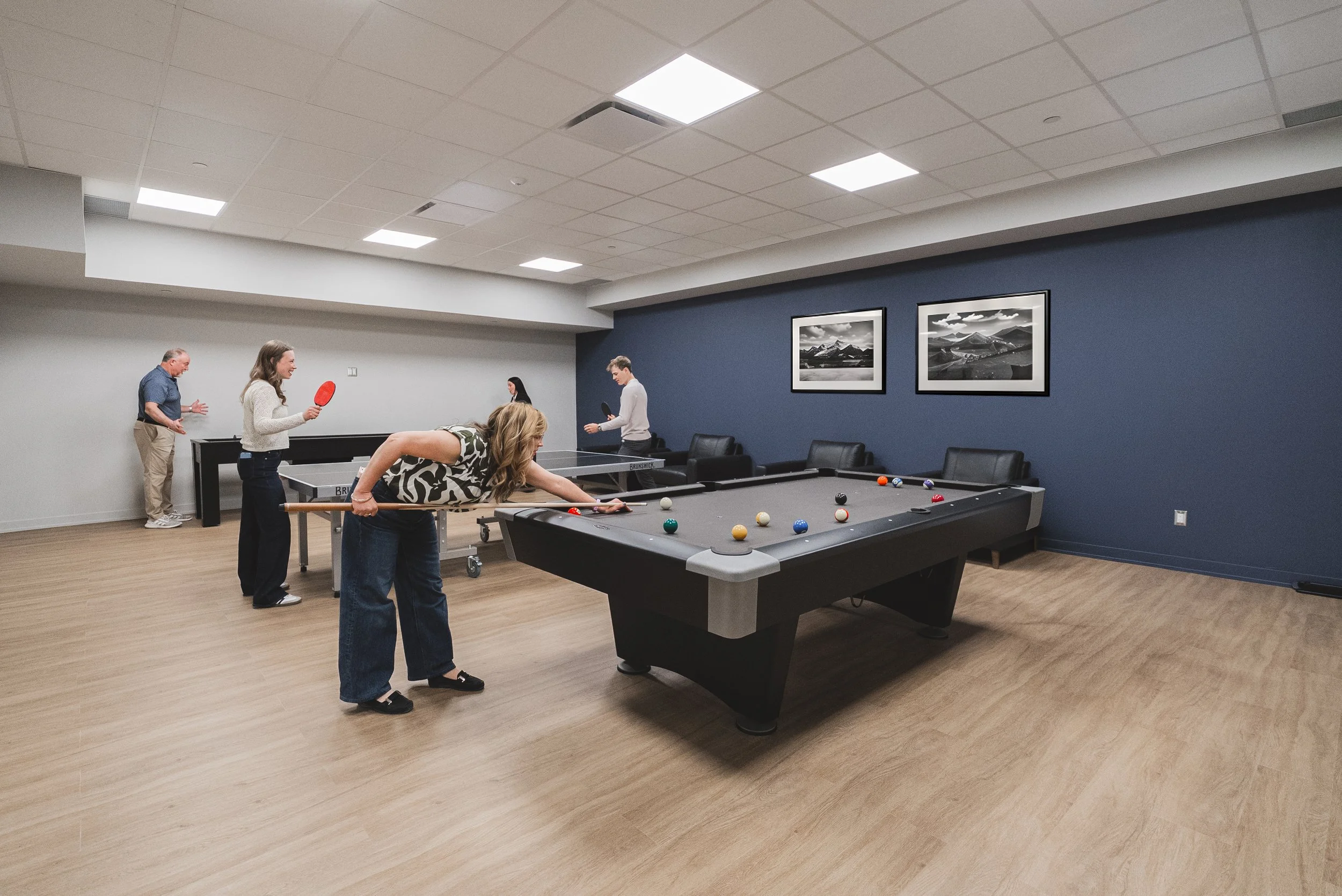 People playing pool in an indoor game room with black and white landscape photos on blue walls and wooden flooring.