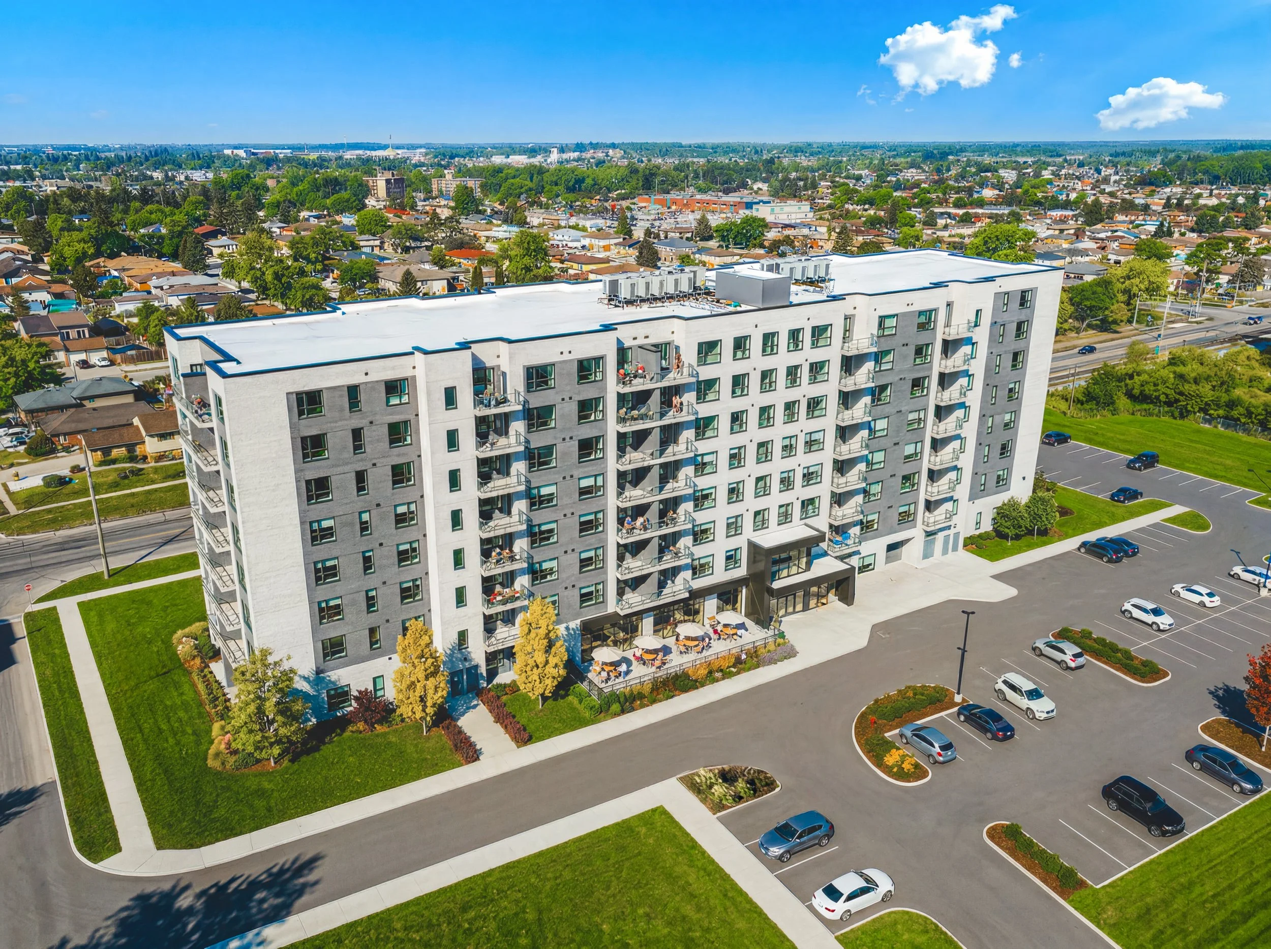 Aerial view of a modern, white multi-story apartment building with balconies, surrounded by green lawns, parking lot with scattered cars, and neighboring residential area under a blue sky with some clouds.