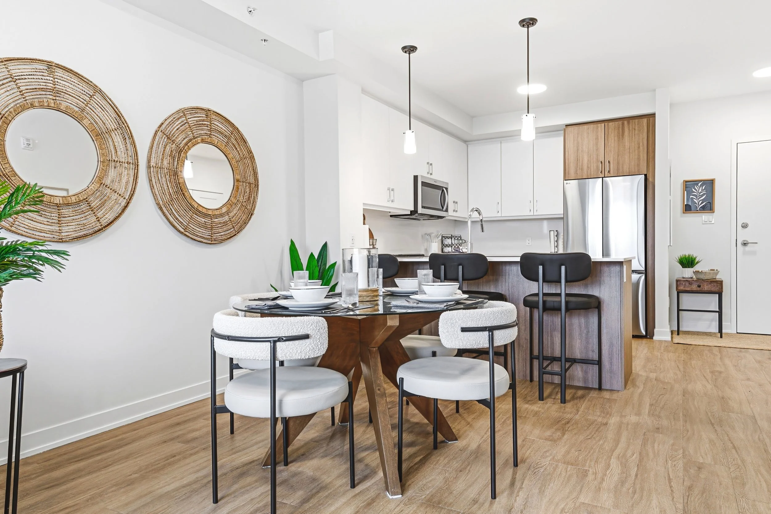 Modern apartment kitchen and dining area with white walls, wooden flooring, and black, white, and wooden furniture. There is a round dining table with four chairs, a kitchen island with two black barstools, and kitchen cabinets with stainless steel a