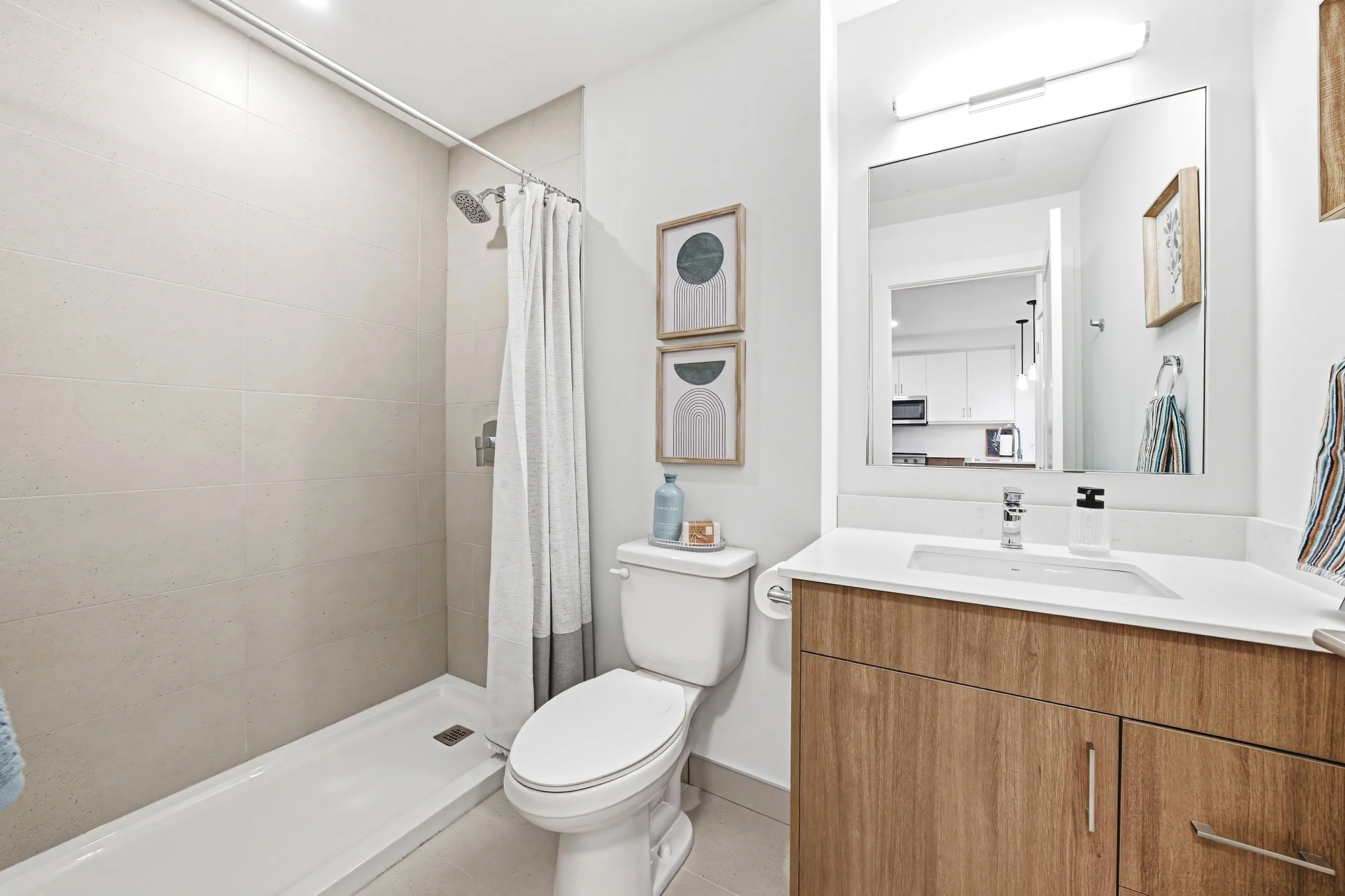 Modern bathroom with walk-in shower with beige tiles, white vanity with wooden cabinet, large mirror, toilet, and minimal decor.