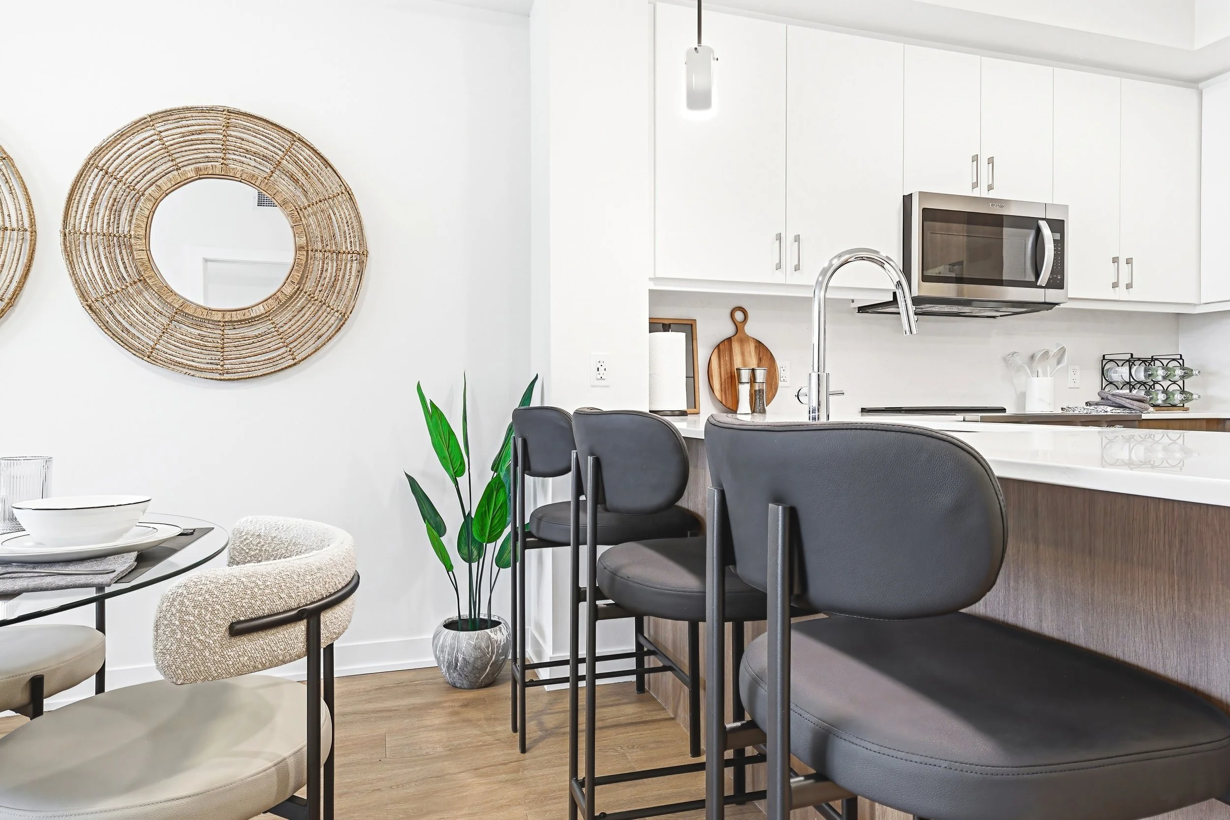 Modern kitchen and dining area with black bar stools, white cabinets, stainless steel microwave, potted plants, decorative wall mirrors, and minimal decor.