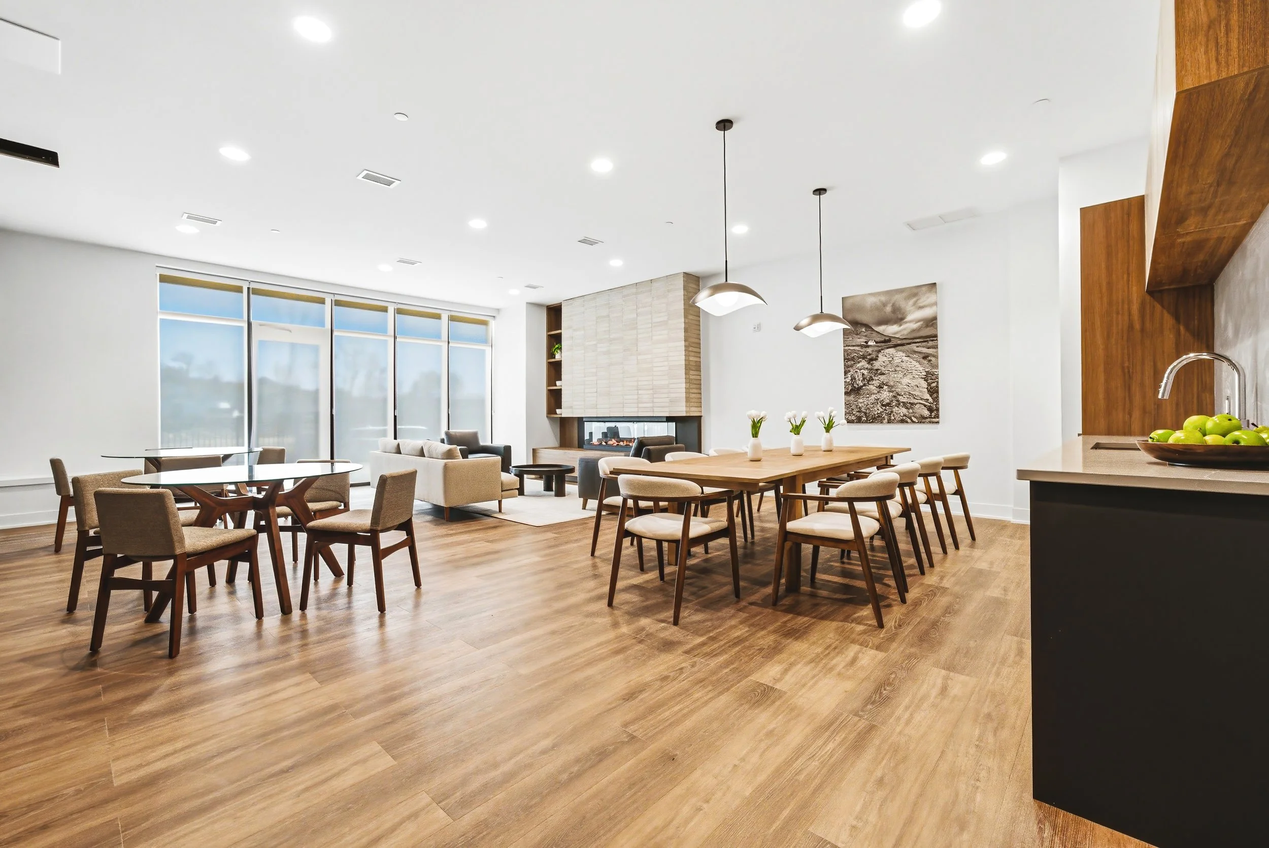 Modern open-concept living and dining area with wooden floors, white walls, large windows, beige and black furniture, a fireplace, and artwork.