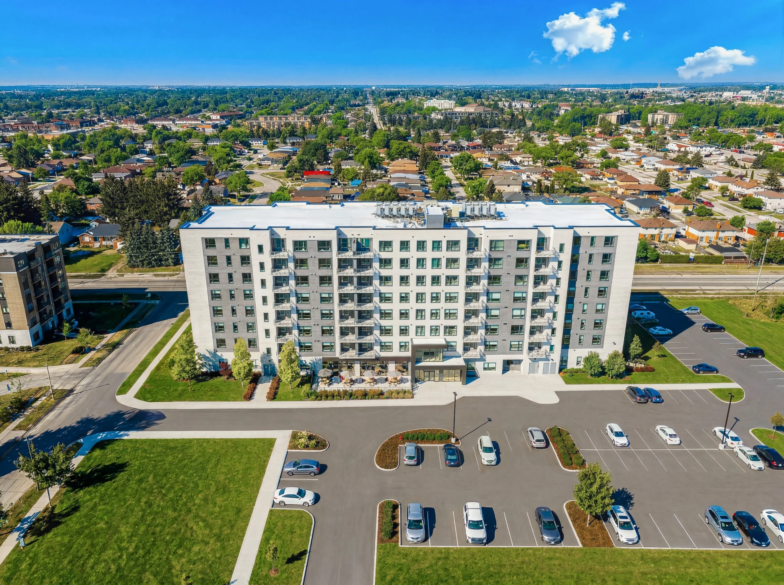 An aerial view of a modern, white multi-story apartment building with parking lots and landscaped green areas in a suburban neighborhood under a bright blue sky.