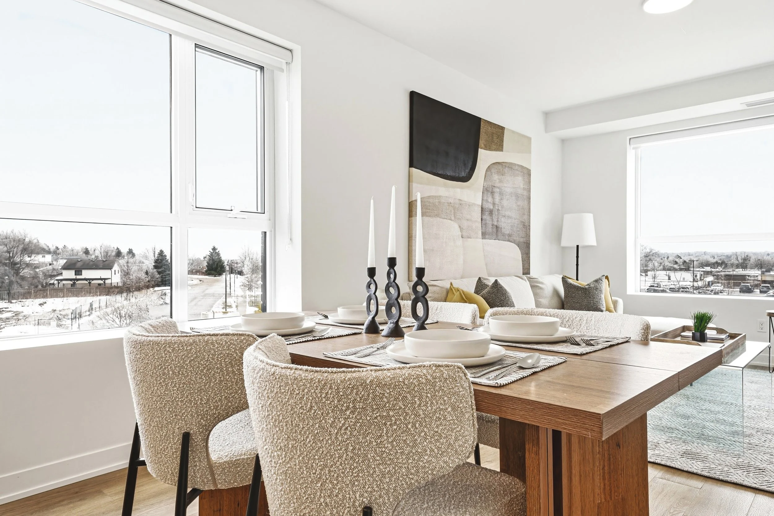 A dining area with a wooden table set with white plates, bowls, utensils, and decorative black candlesticks with white candles. Beige upholstered chairs surround the table. Behind, a white sofa with gray and yellow pillows is visible, with a large ab