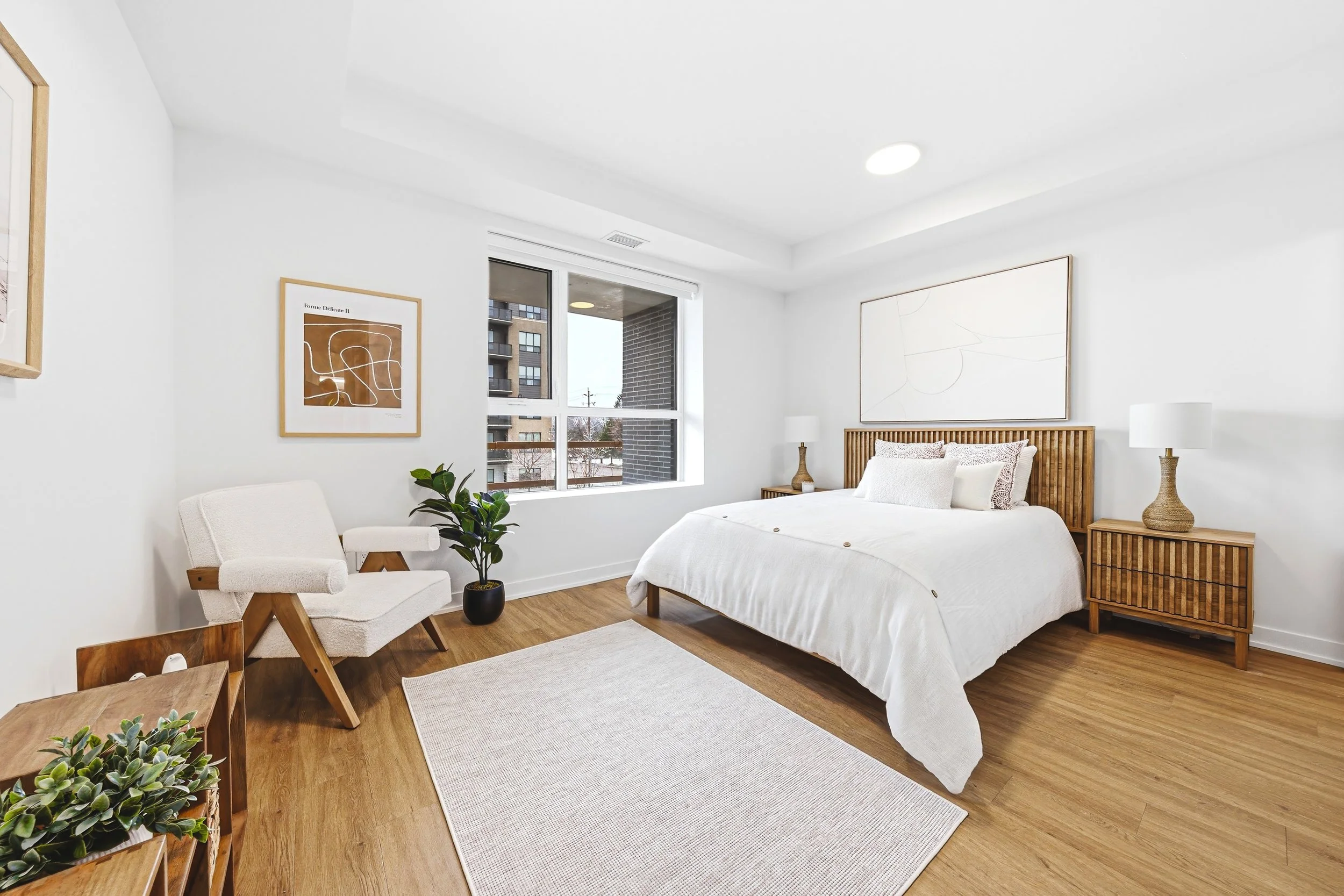 Modern bedroom with white walls, wooden furniture, a bed with white bedding, bedside tables with lamps, a chair, a potted plant, framed artwork, and a large window with an outside view.