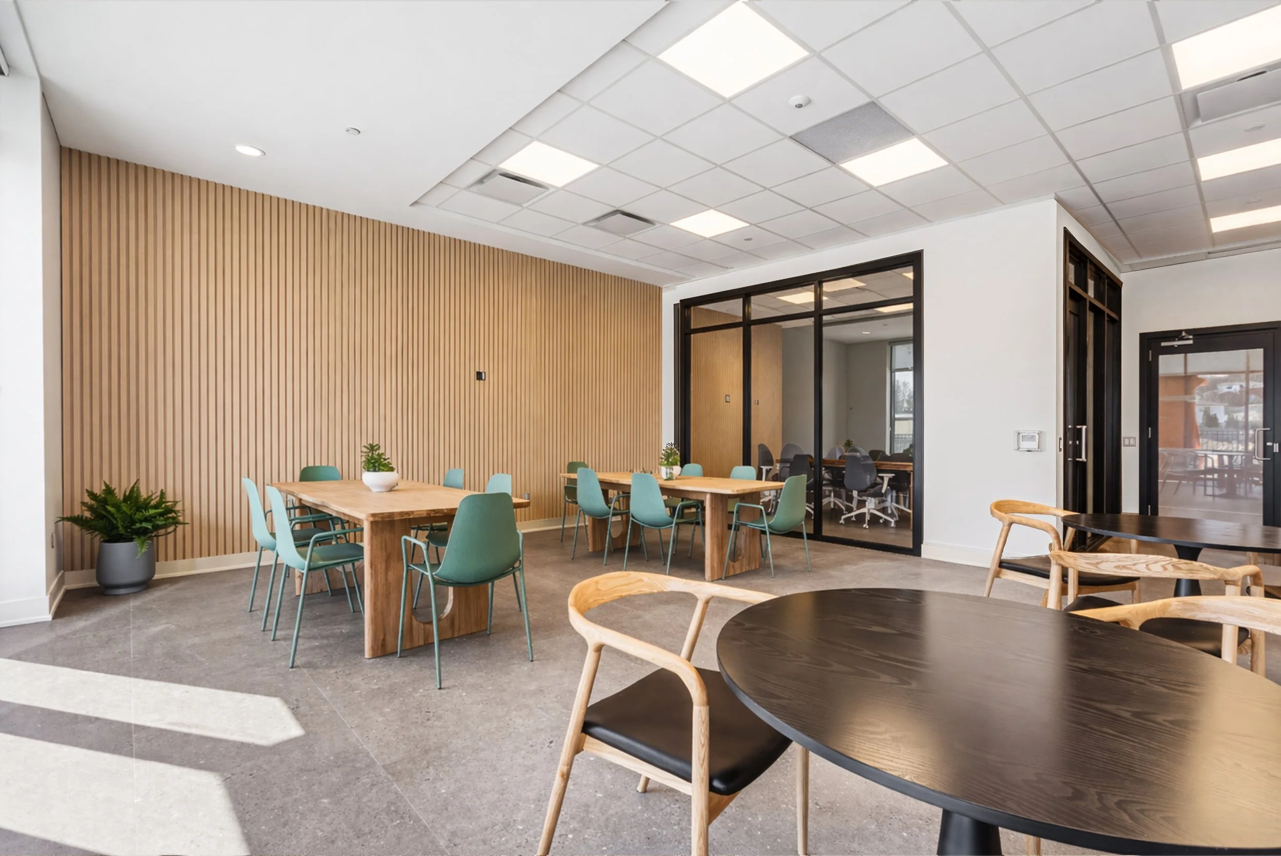 Modern office break room with round black tables and wooden chairs, a wooden wall with vertical slats, and a glass-walled private office area.
