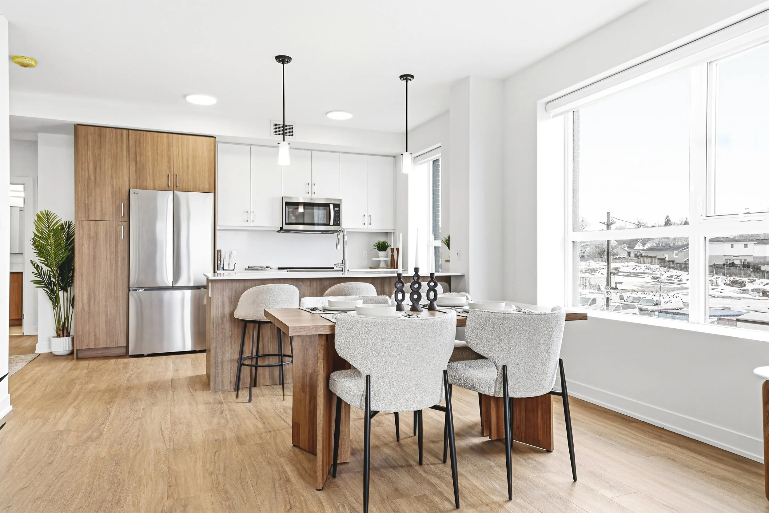 Modern kitchen with a wooden dining table set for a meal, surrounded by four light gray upholstered chairs, with large windows providing natural light and a view of a snowy outdoor scene.