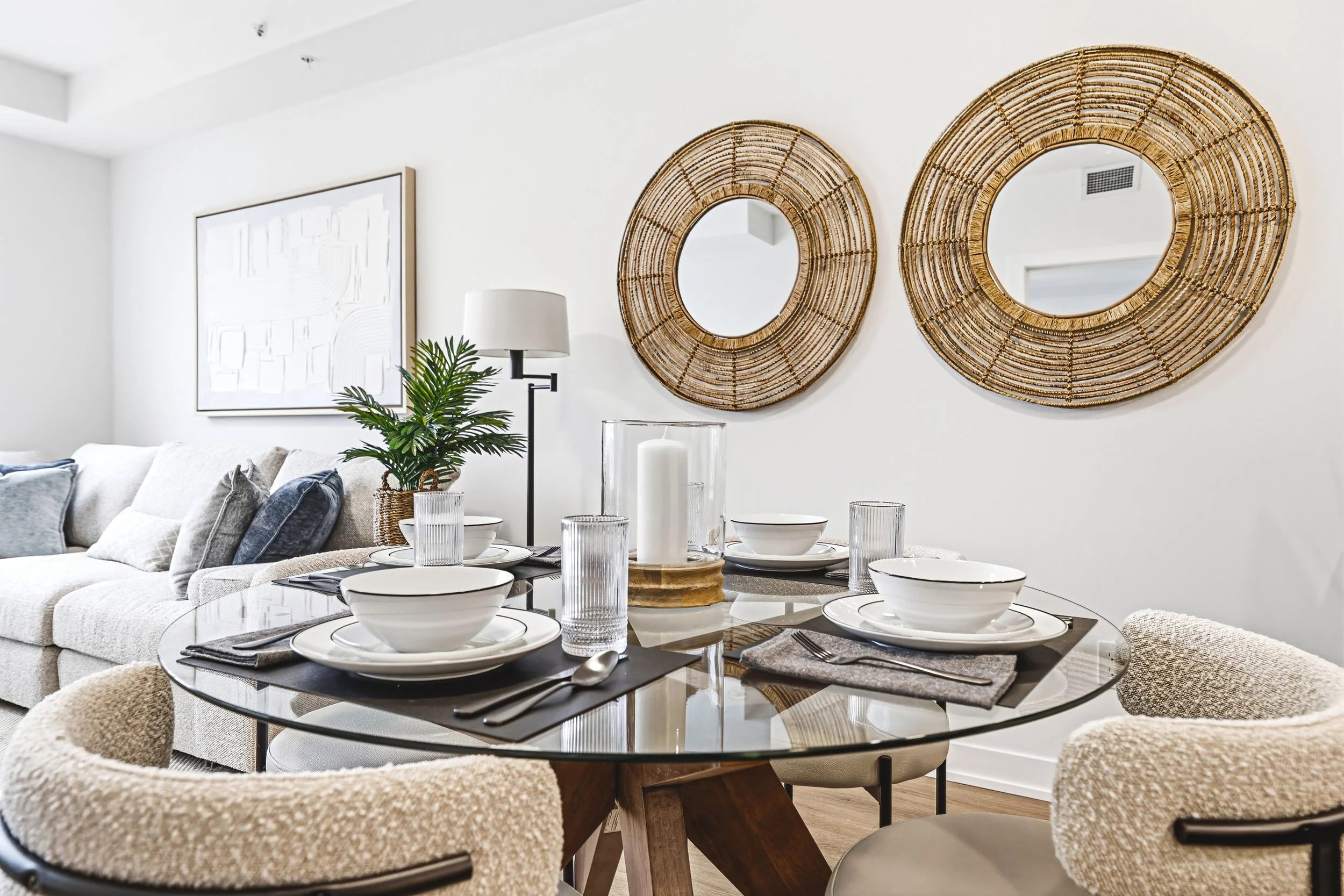 Dining table set with white bowls, plates, and glasses, surrounded by beige chairs, in a modern living space with two round woven mirrors and a potted plant on a white wall.