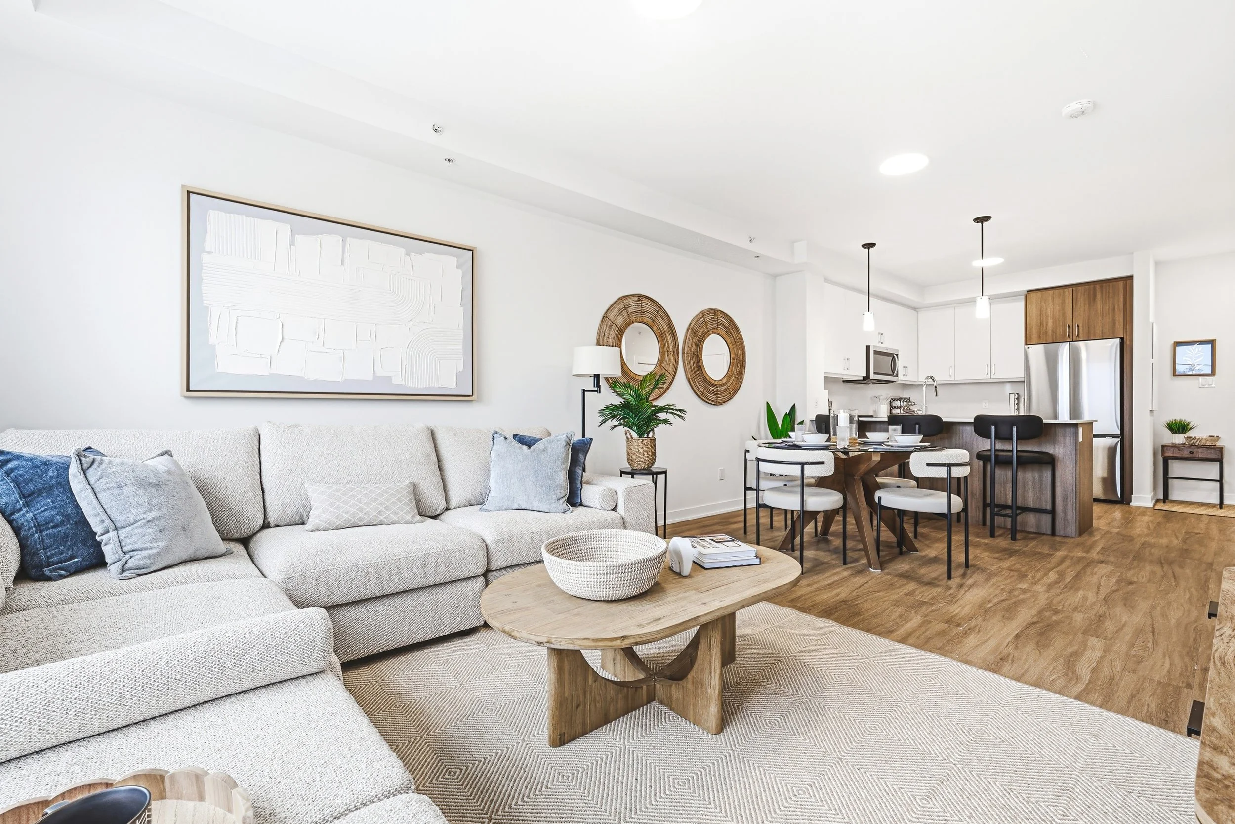 Modern living room and dining area with white walls, beige sofa with cushions, wooden table, and a kitchen with stainless steel appliances.