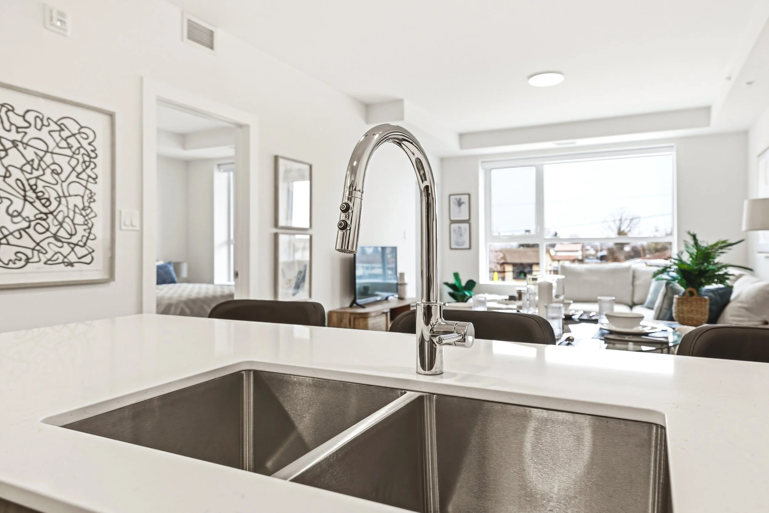 Modern kitchen faucet with a double-basin stainless steel sink on a white countertop in a bright open living space.
