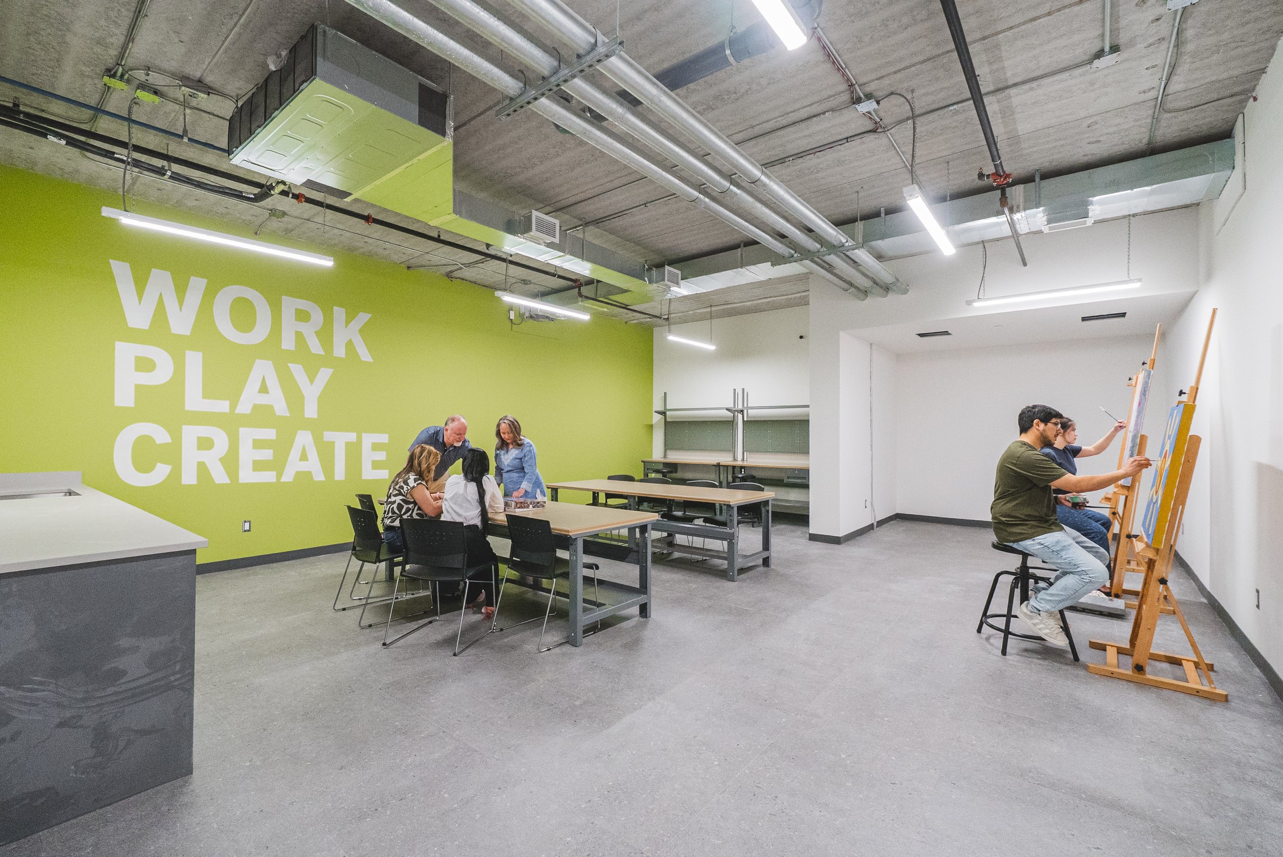People drawing on canvases in an industrial-style art studio with a lime green wall that says "WORK PLAY CREATE".