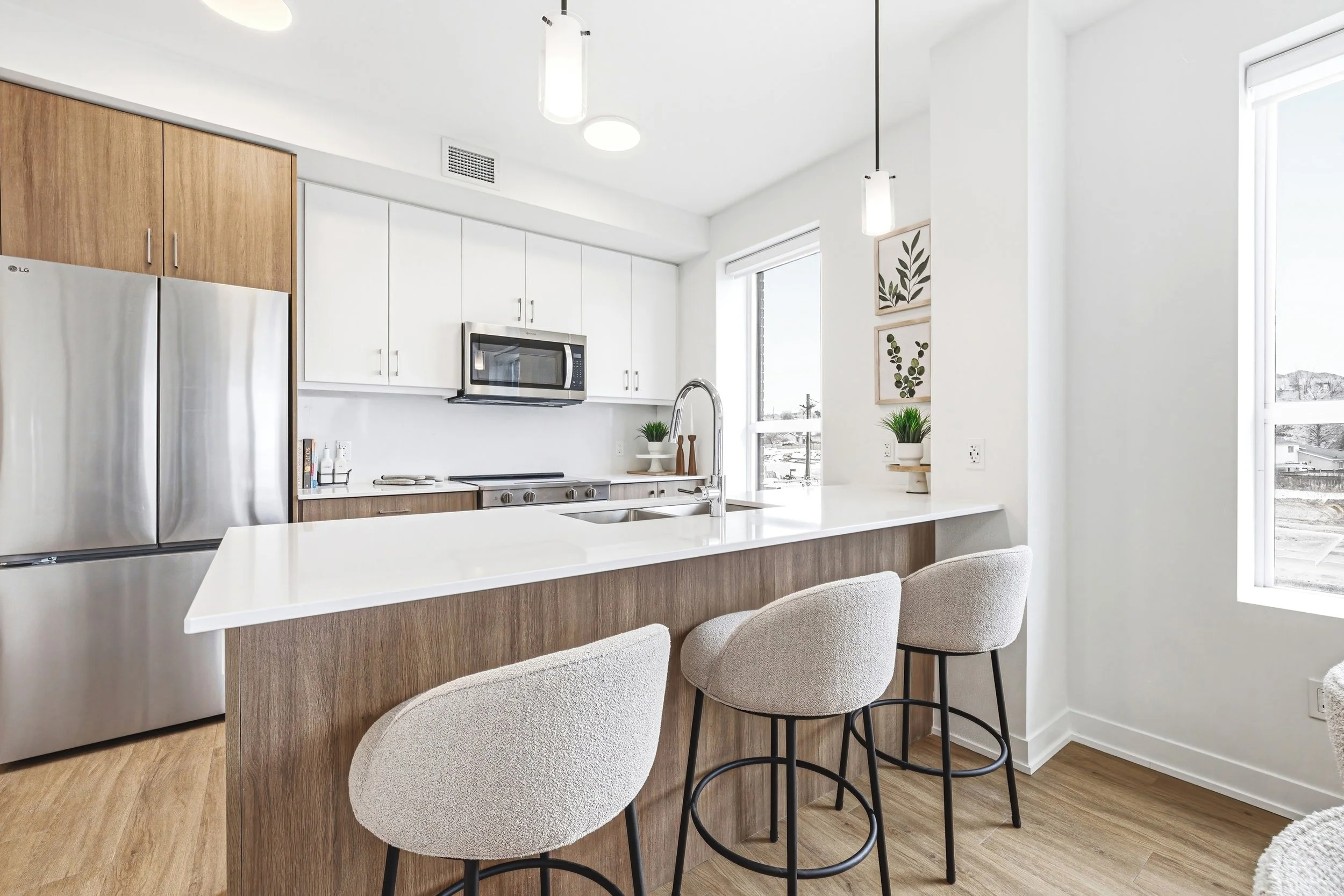Modern kitchen with white and wood cabinetry, stainless steel appliances, a white countertop island with three beige chairs, and decorated with green plants and artwork near the window.