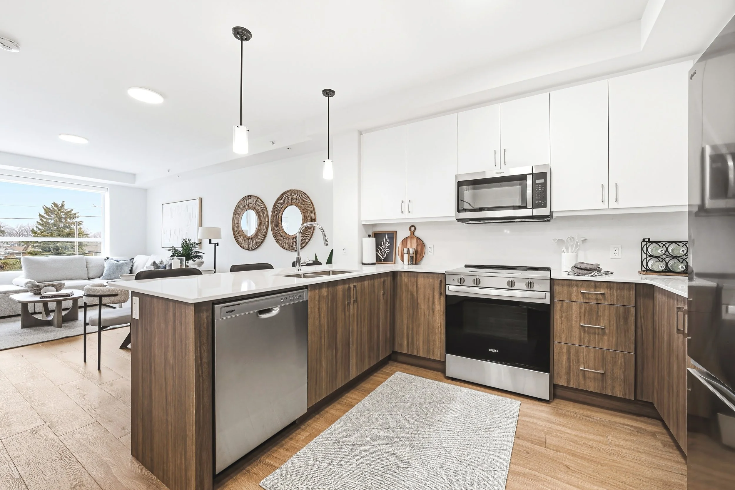 Modern kitchen with white cabinets, stainless steel appliances, wood lower cabinets, and a white countertop island. Adjacent living area with sofa, chairs, and large window.