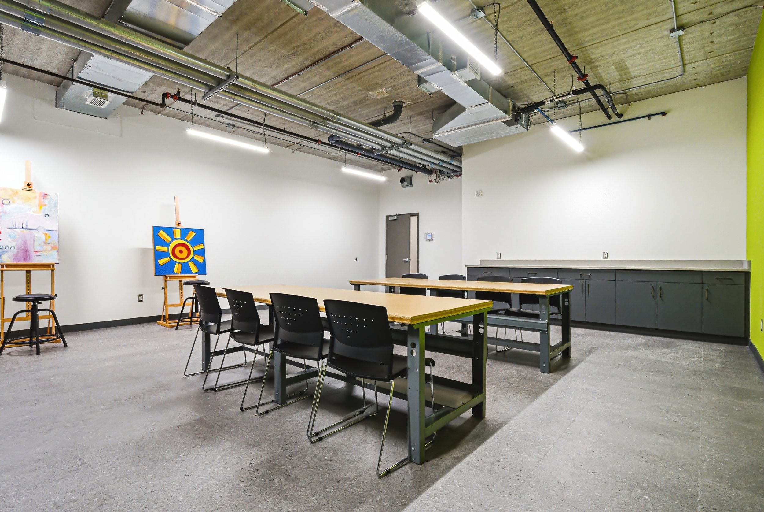 Interior of a modern meeting or classroom space with tables, chairs, and exposed ceiling pipes and ductwork.
