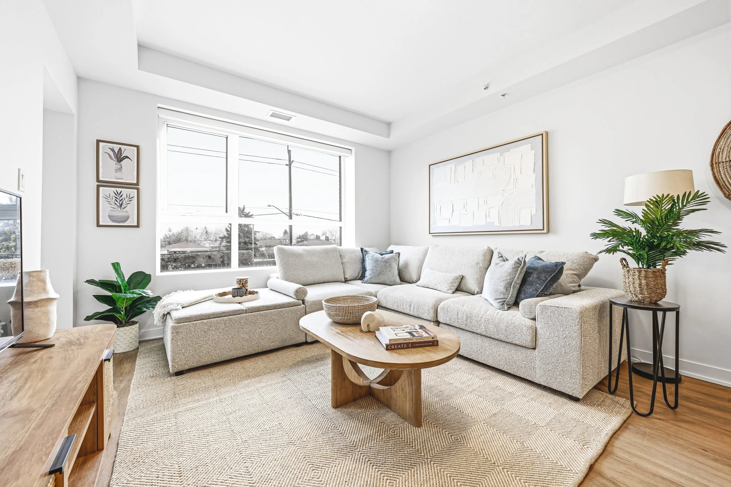 Bright living room with white walls, large window with blinds, beige sectional couch with pillows, wooden coffee table with books and decorative items, flat-screen TV, and potted plants.