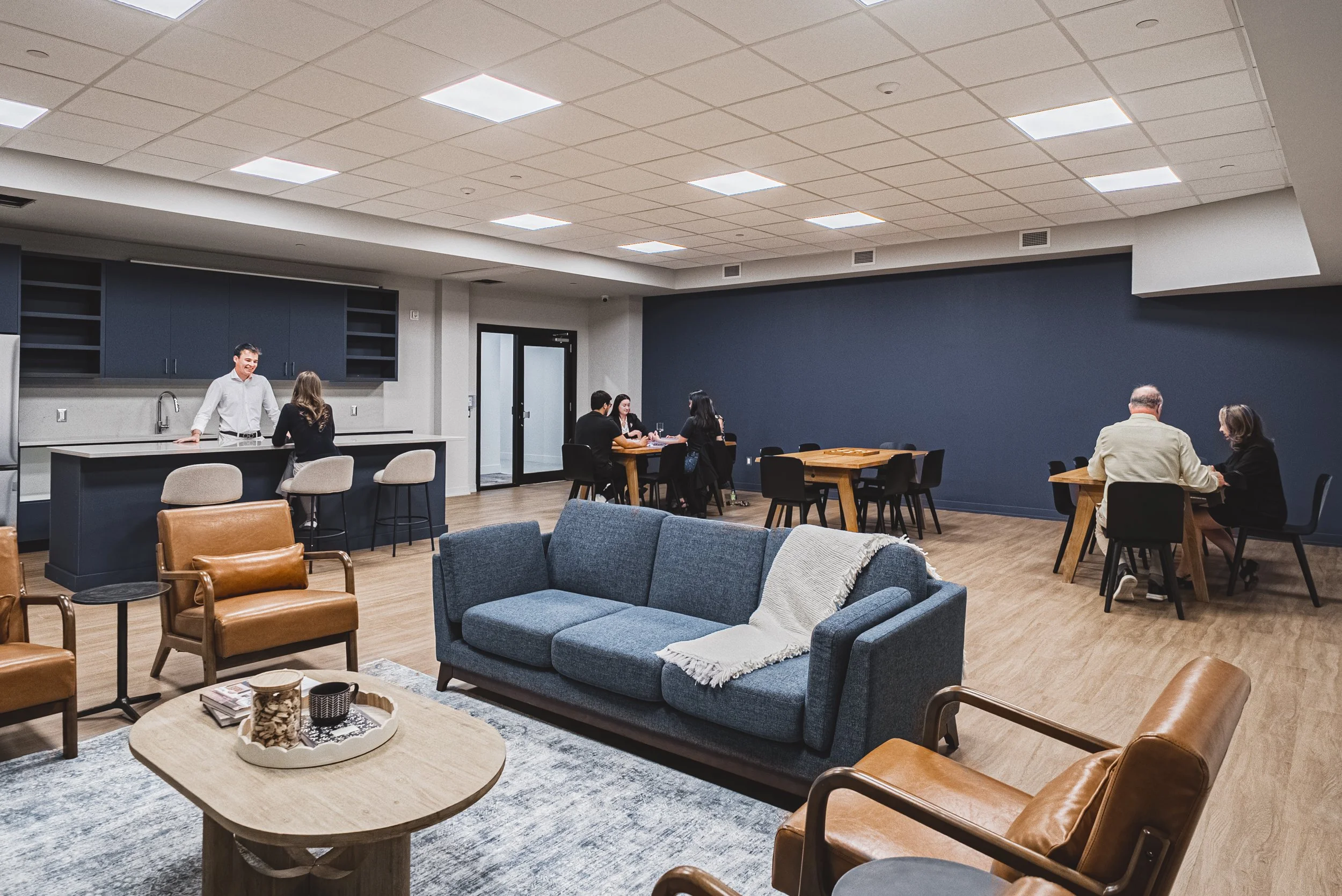 A spacious indoor lounge or break room with modern furniture, including a sofa, armchairs, and tables. Several people are sitting and talking, and one person stands by a kitchen counter. The room has drop ceiling tiles, bright lighting, and a dark accent wall.