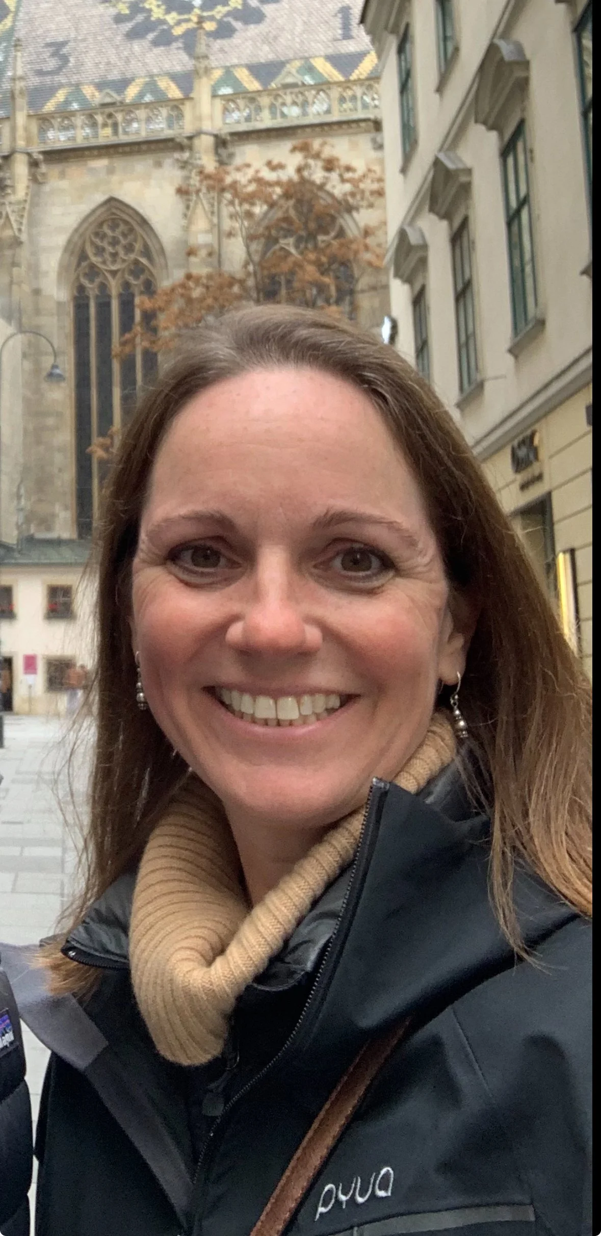 A woman smiling, wearing a beige scarf, earrings, and a black jacket. Behind her is a historic building, possibly a cathedral, with stained glass windows and detailed stone architecture.