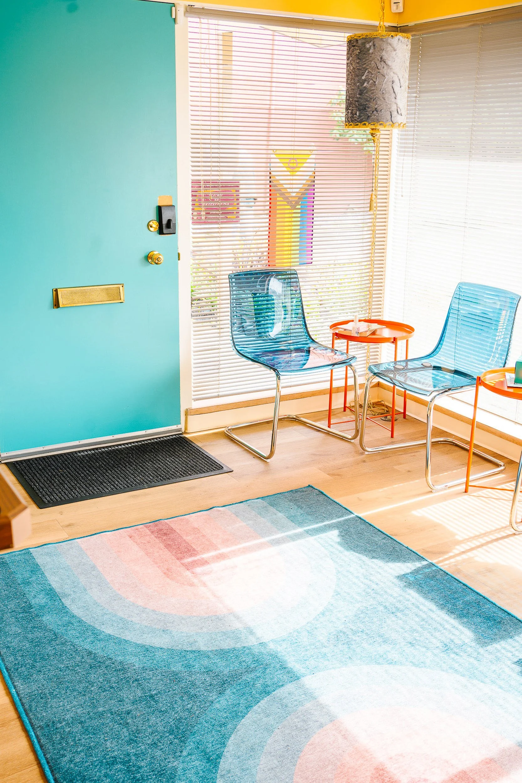 A colorful rainbow-patterned area rug on a wooden floor in a room with a teal door, blue chairs, orange tables, and window blinds letting in sunlight.