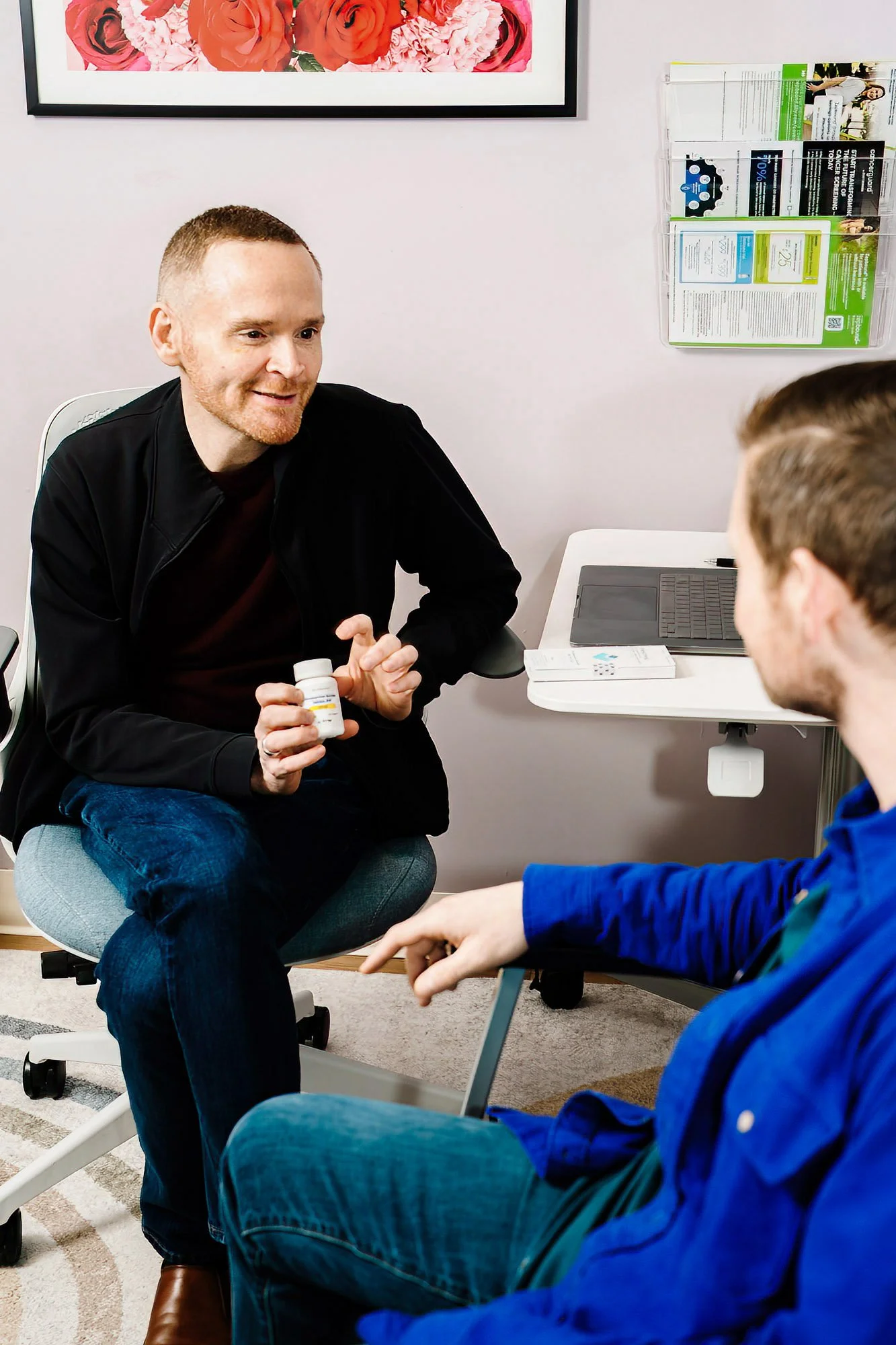 A man with red hair and a beard, wearing a black jacket, is sitting in a chair and holding a white medication bottle. He is talking to a younger person, who is wearing a blue jacket. They are in a medical office with a white wall, framed floral artwork, and a brochure holder on the wall.