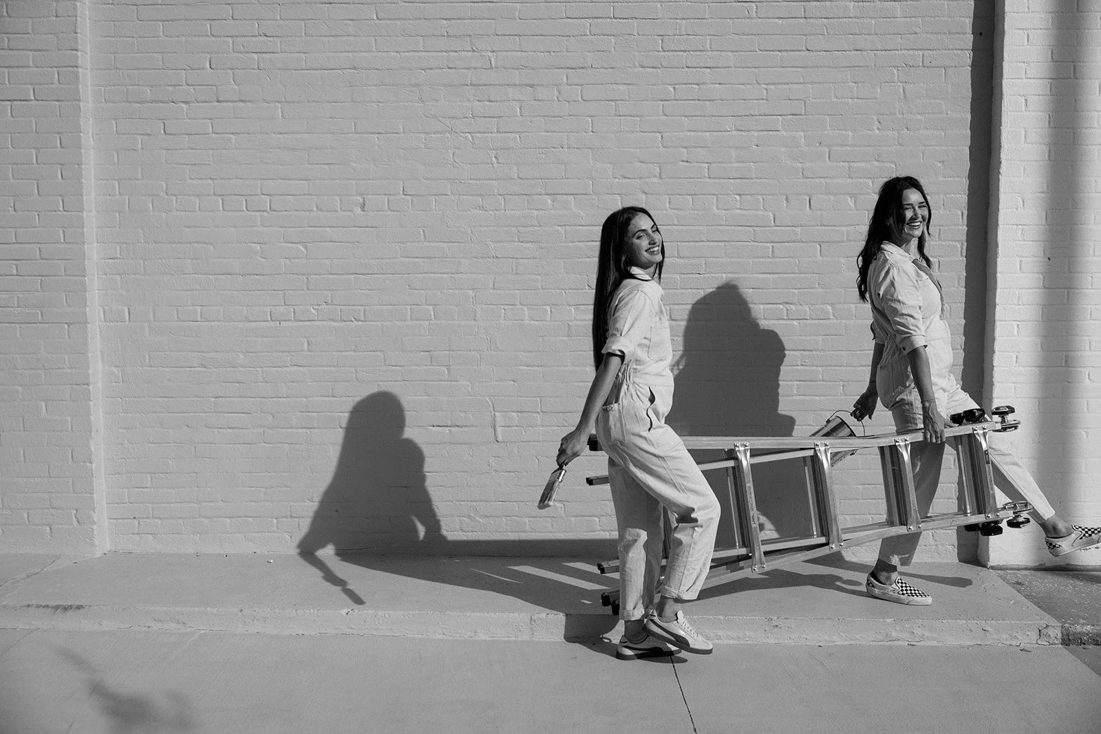 Two women in casual clothing carrying a ladder together while walking along a sidewalk next to a brick wall, smiling and enjoying each other's company.