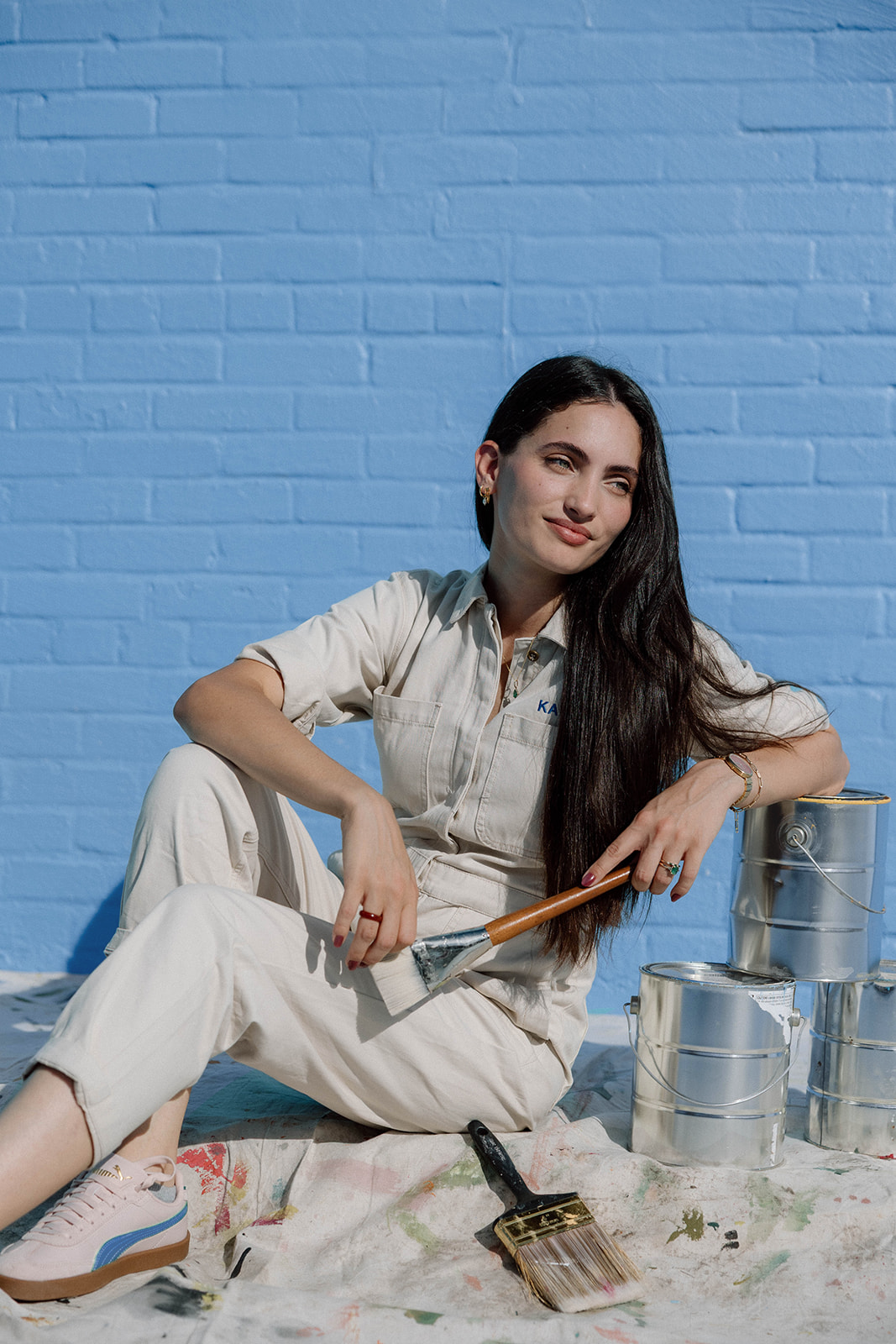 A young woman with long dark hair sitting on a cloth surrounded by paint cans, holding a paintbrush, with a painted blue brick wall in the background.