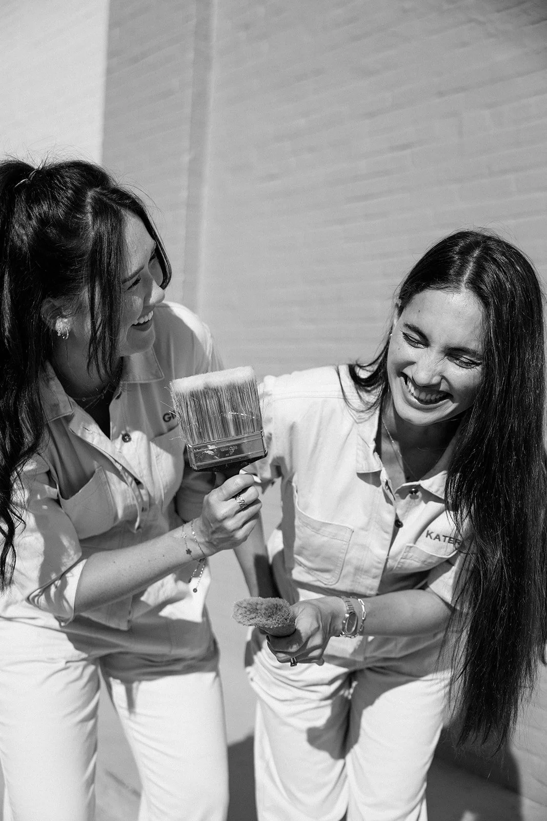 Two women in work uniforms laughing and smiling at each other, one holding a paintbrush and the other holding a sponge.
