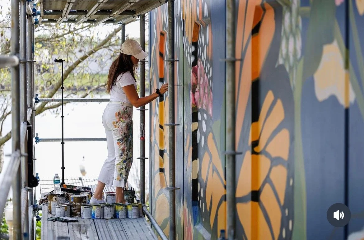 A woman painting a colorful mural on a wall outdoors, standing on a scaffold with paint cans nearby.