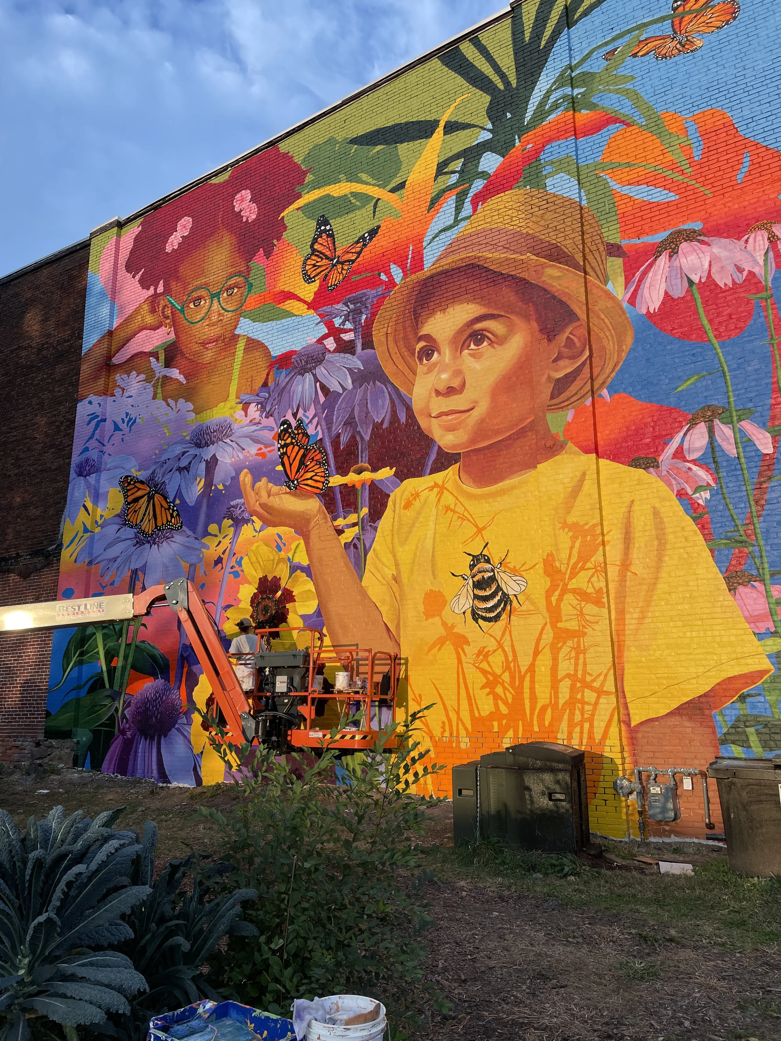 A large colorful mural on a building wall depicting two children, a boy in a yellow shirt and hat, and a girl with glasses and pink hair. The mural features flowers, butterflies, and a bee, with vibrant colors and detailed artwork. Mural painting is in progress, with artists working on it and an orange lift in front.