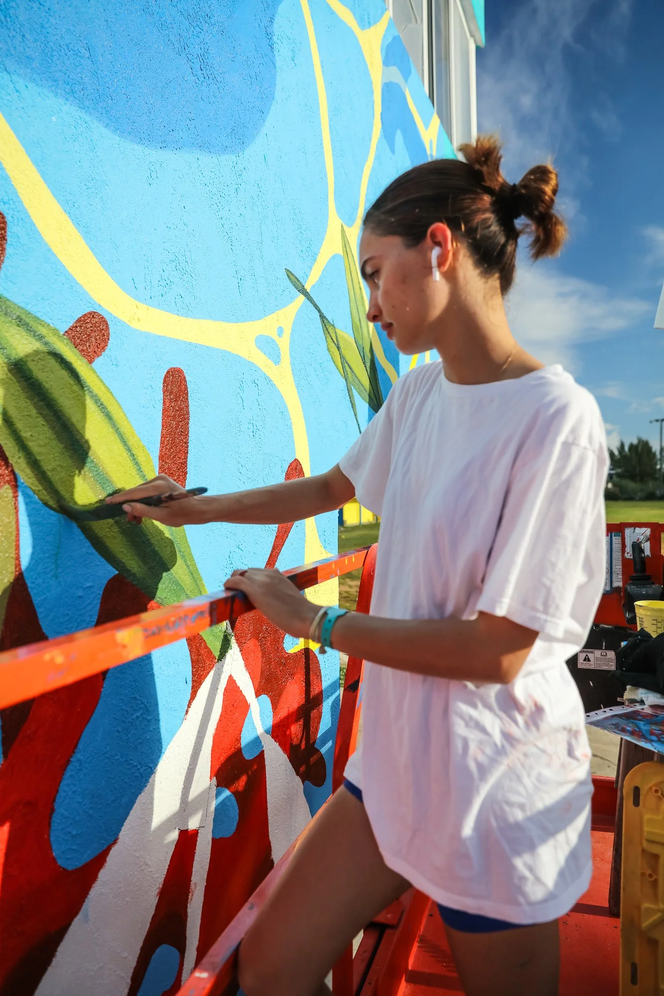 A young woman is painting a colorful mural on an exterior wall using a paintbrush. She is standing on a scaffold and is wearing a white T-shirt, navy shorts, and wireless earbuds. The mural features abstract shapes and bright colors, and the sky is clear with some clouds visible.