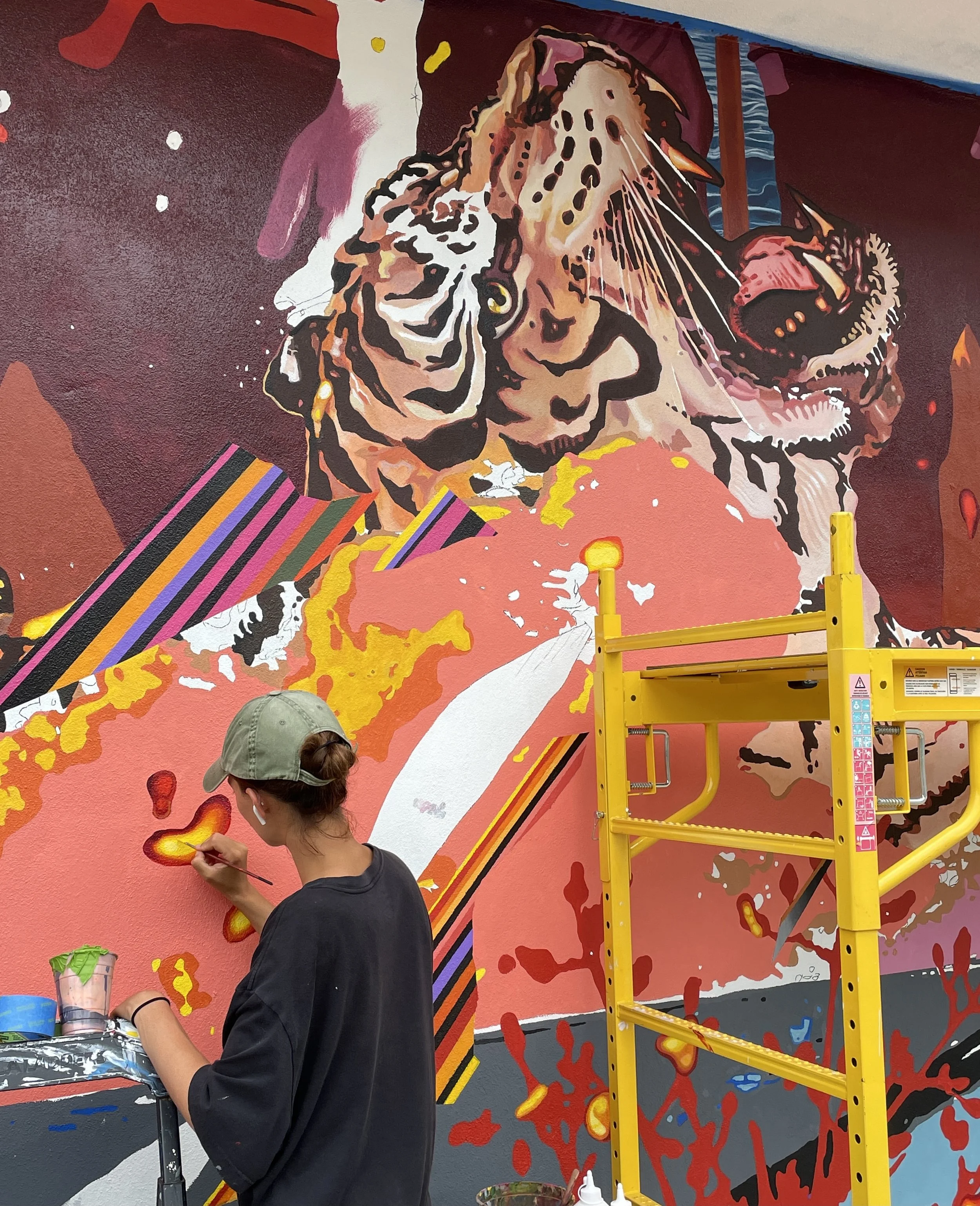 A woman painting a large, colorful mural of a tiger's face on a wall, using bright colors and bold patterns, with art supplies and a yellow ladder nearby.