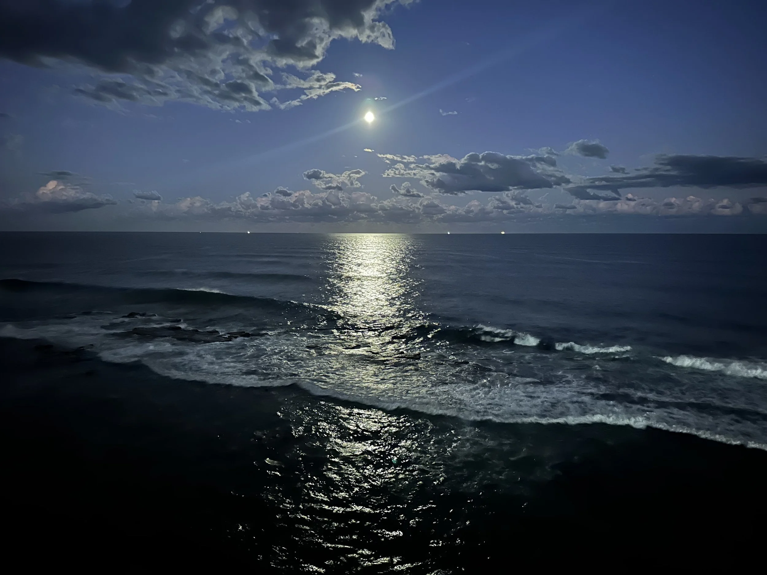 The image shows a night view over the ocean with a full moon shining brightly in a partly cloudy sky, reflecting on the water surface, and waves in the foreground.