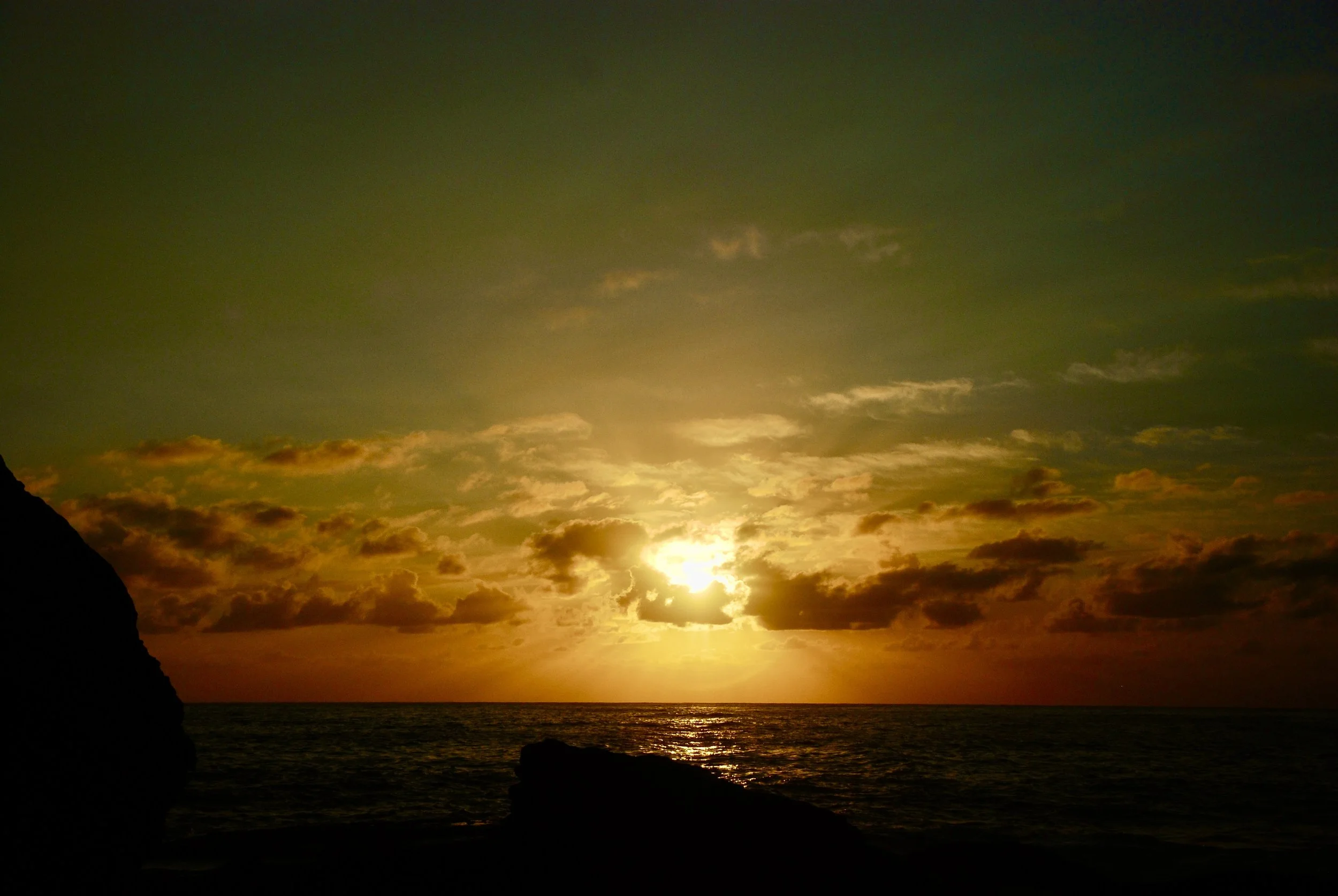 A sunset over the ocean with orange and golden clouds, silhouette of a rocky coastline on the left.