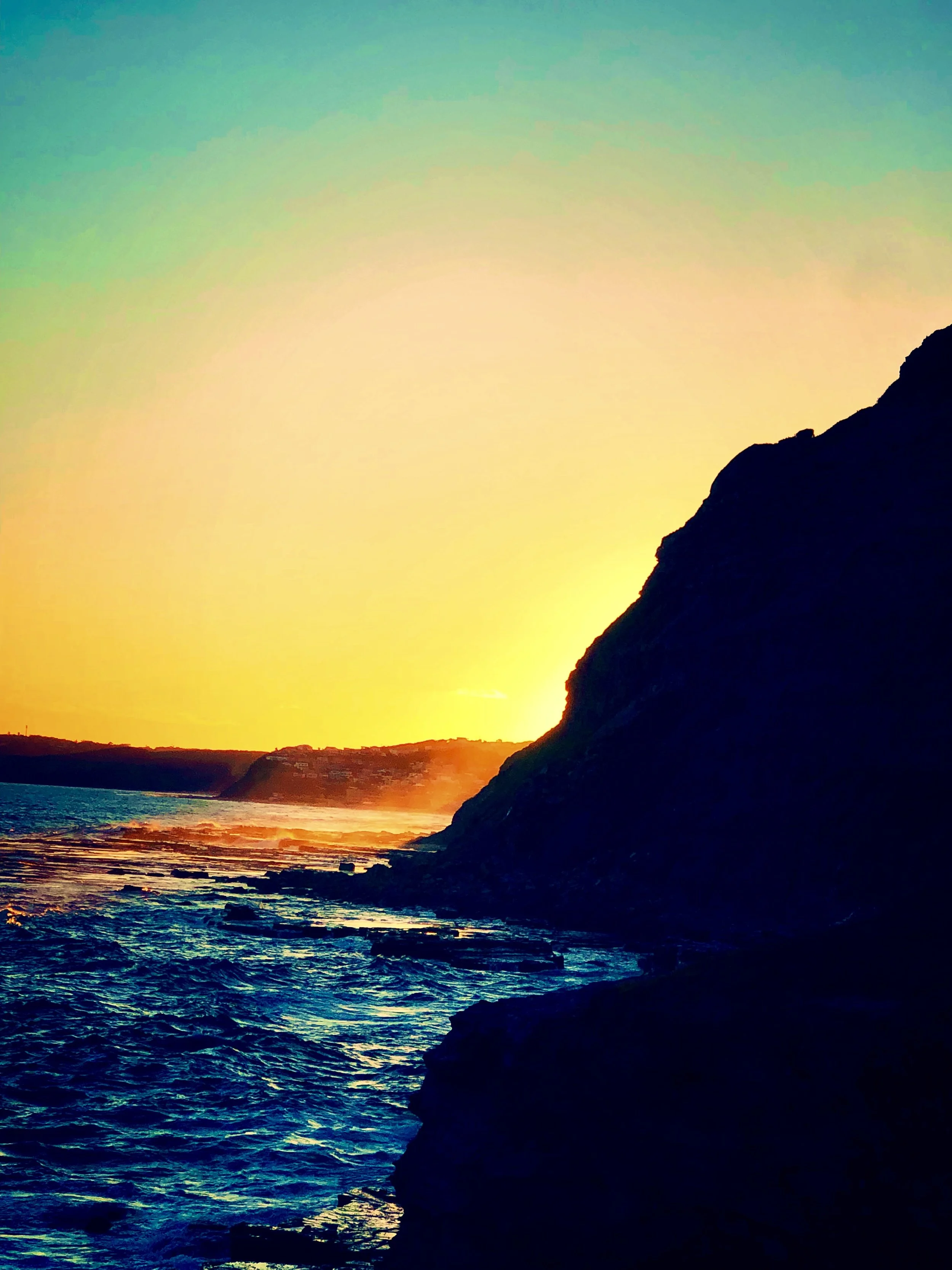 Sunset over the ocean with a silhouette of a rocky cliff on the right side, calm waters in the foreground, and a colorful sky with shades of yellow, orange, and green.