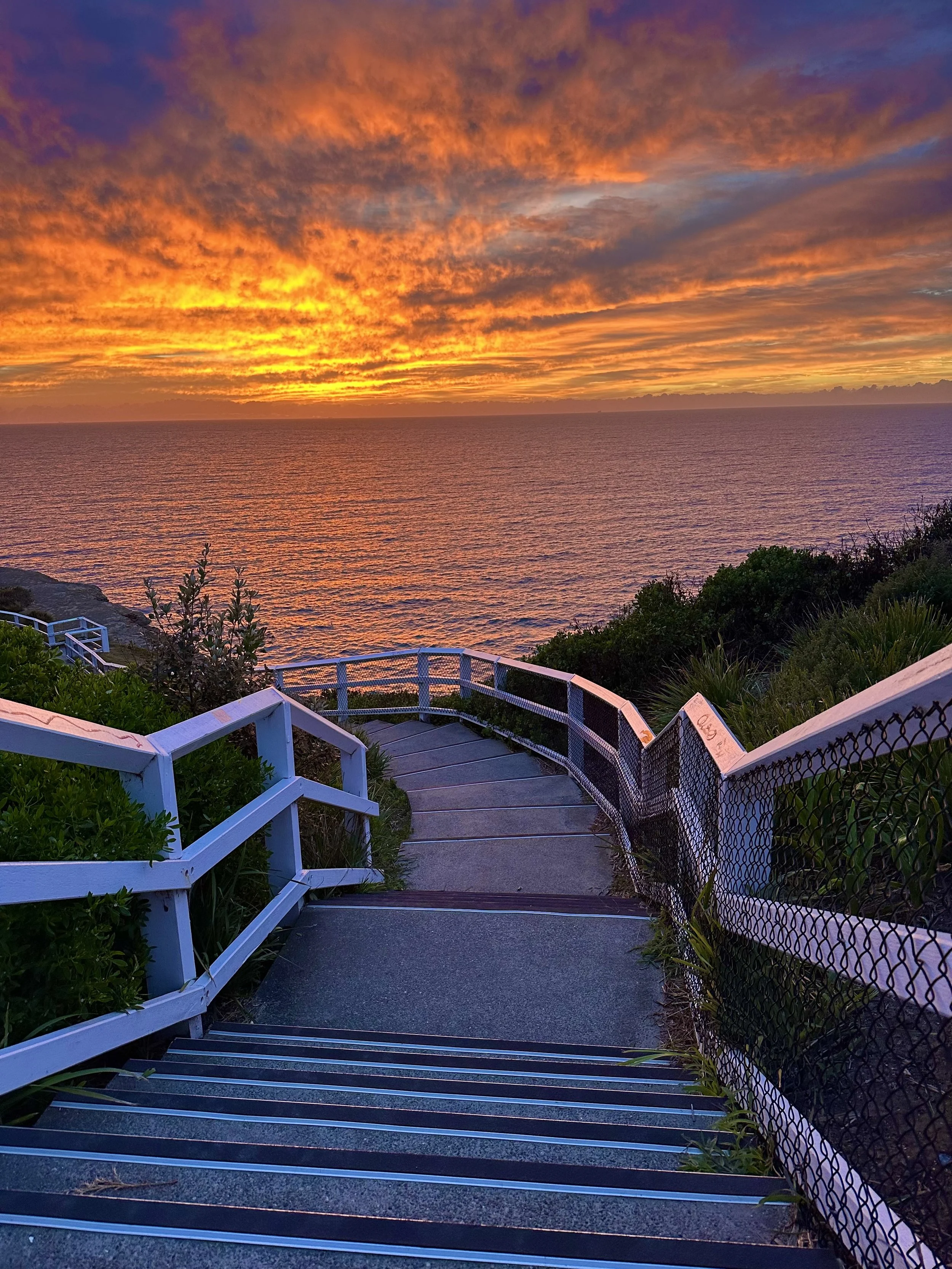 Sunset over the ocean with colorful clouds in the sky, view from a seaside walkway with white railing and greenery.