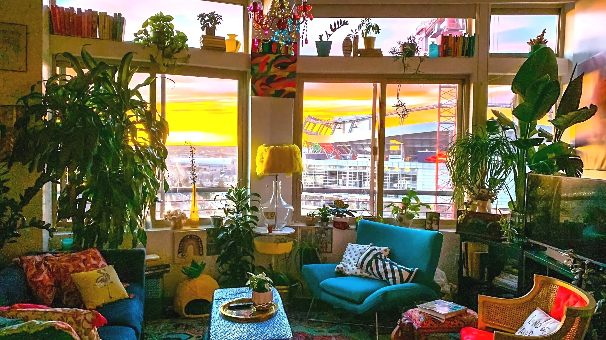 A cozy living room with large windows showing a cityscape with a bridge and colorful sky at sunset. The room is decorated with numerous green plants, colorful pillows, a yellow table lamp, and various art pieces and books.