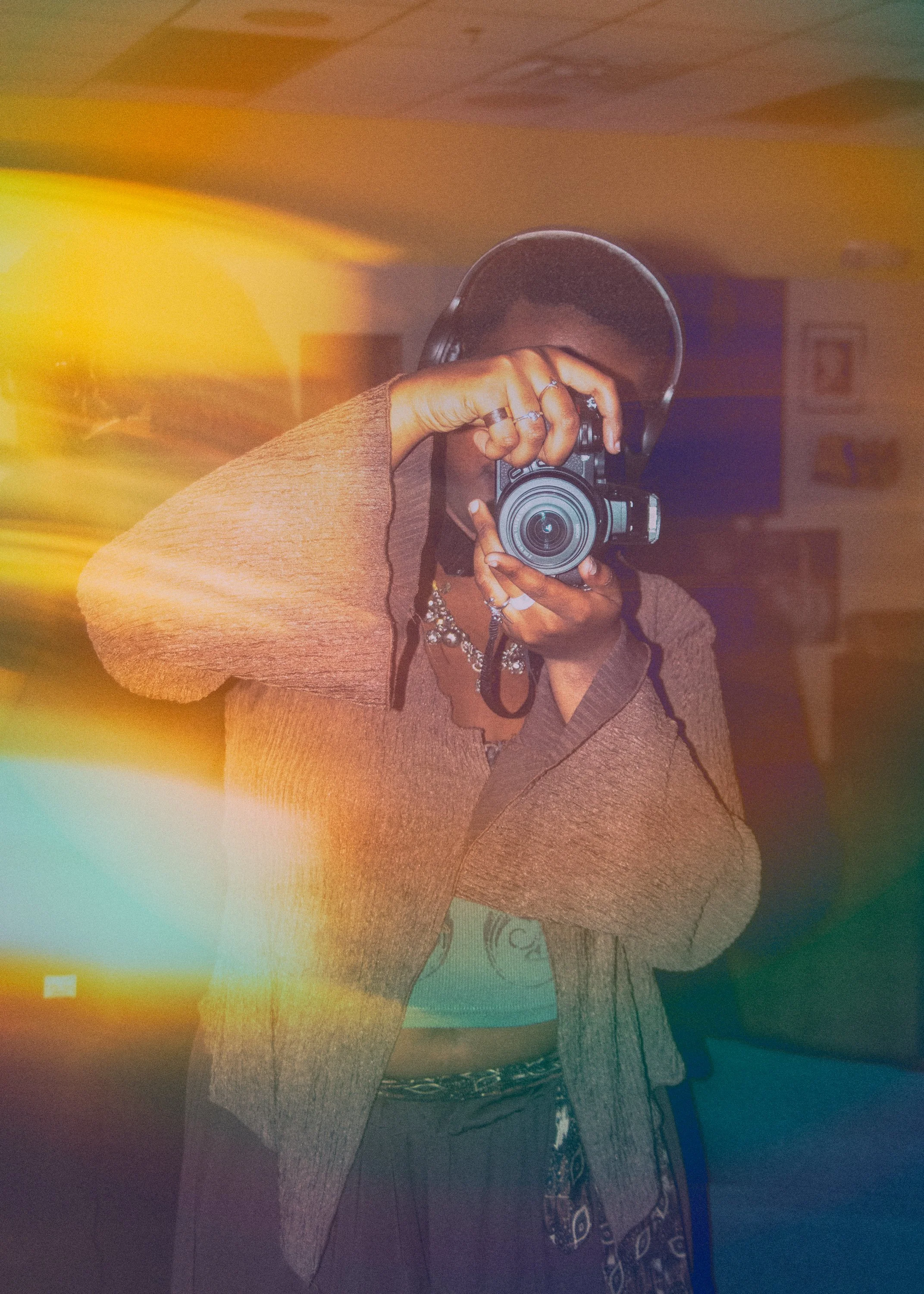 A person taking a selfie with a camera, wearing a brown sweater, ringed fingers, and a hat inside a room with blurred background. Light effects with rainbow-like colors are visible in the image.