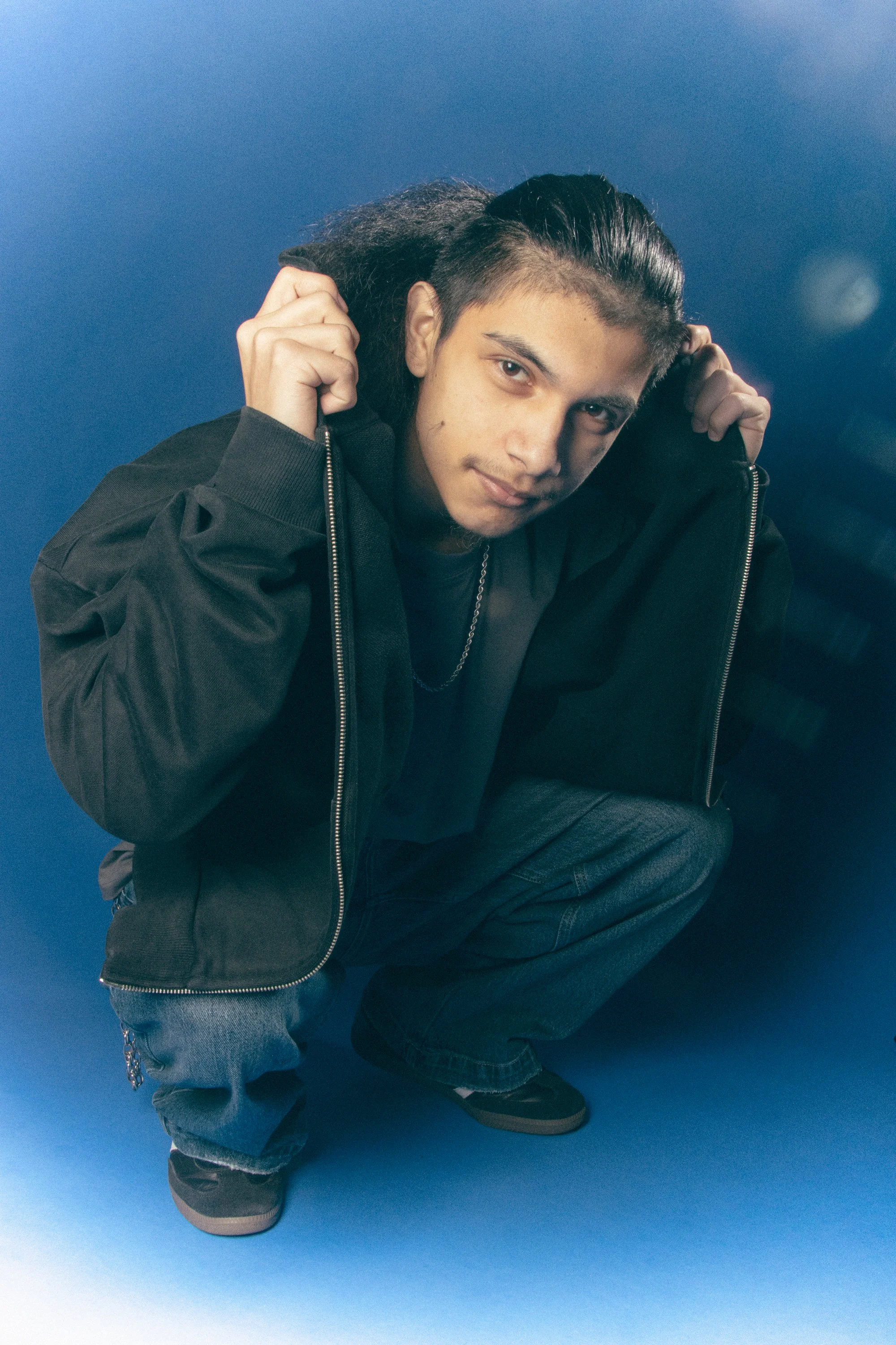 Young man with black hair styled in a pompadour, squatting on a blue background, wearing a black jacket, gray pants, and black sneakers, holding the collar of his jacket.