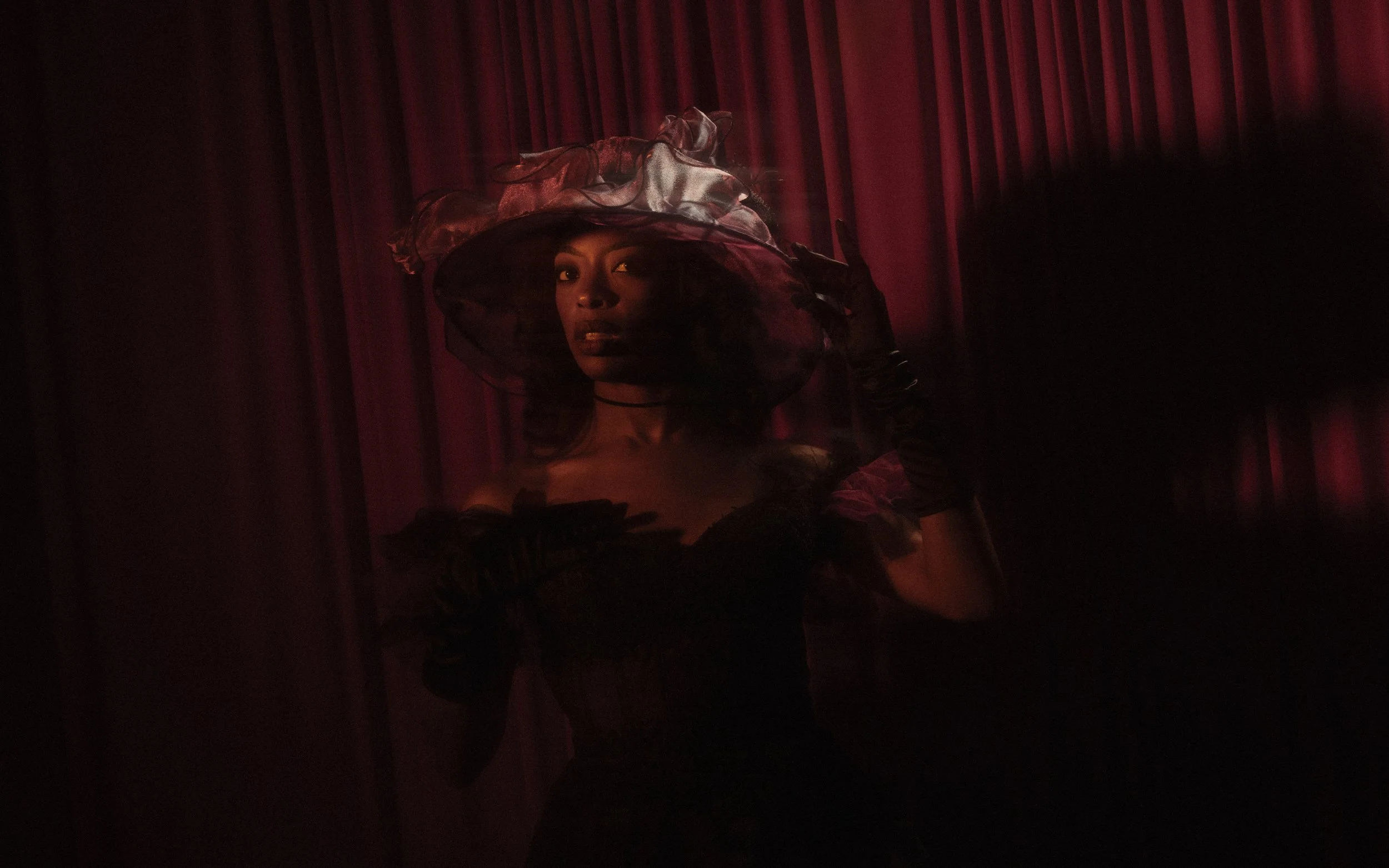 A woman dressed in dark, elaborate clothing with a large, sheer, and ruffled hat poses in front of red stage curtains, partially obscured by shadows.
