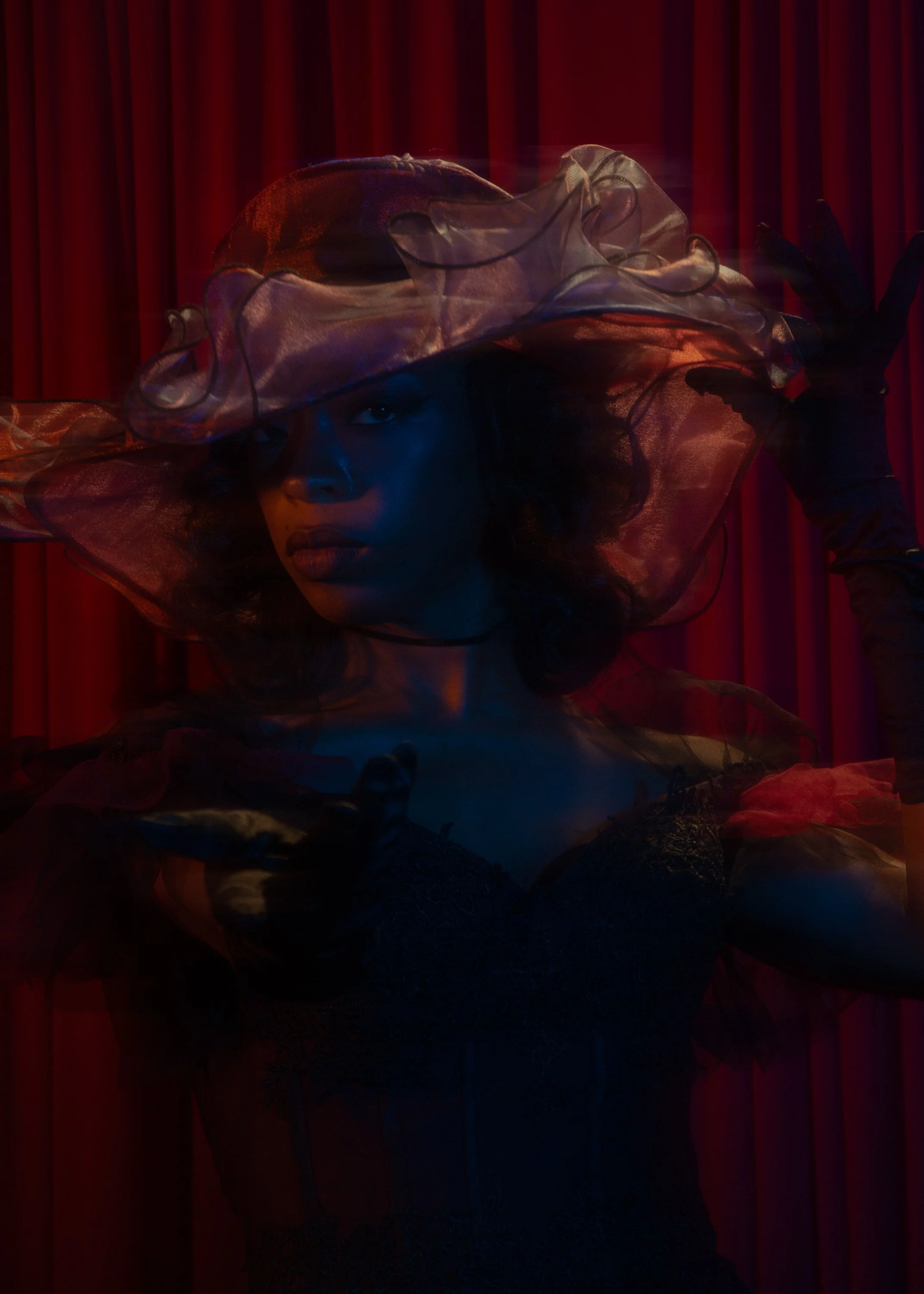 A woman wearing a large, sheer, wide-brimmed hat and dark gloves poses in front of red curtains, illuminated by blue and red lighting.
