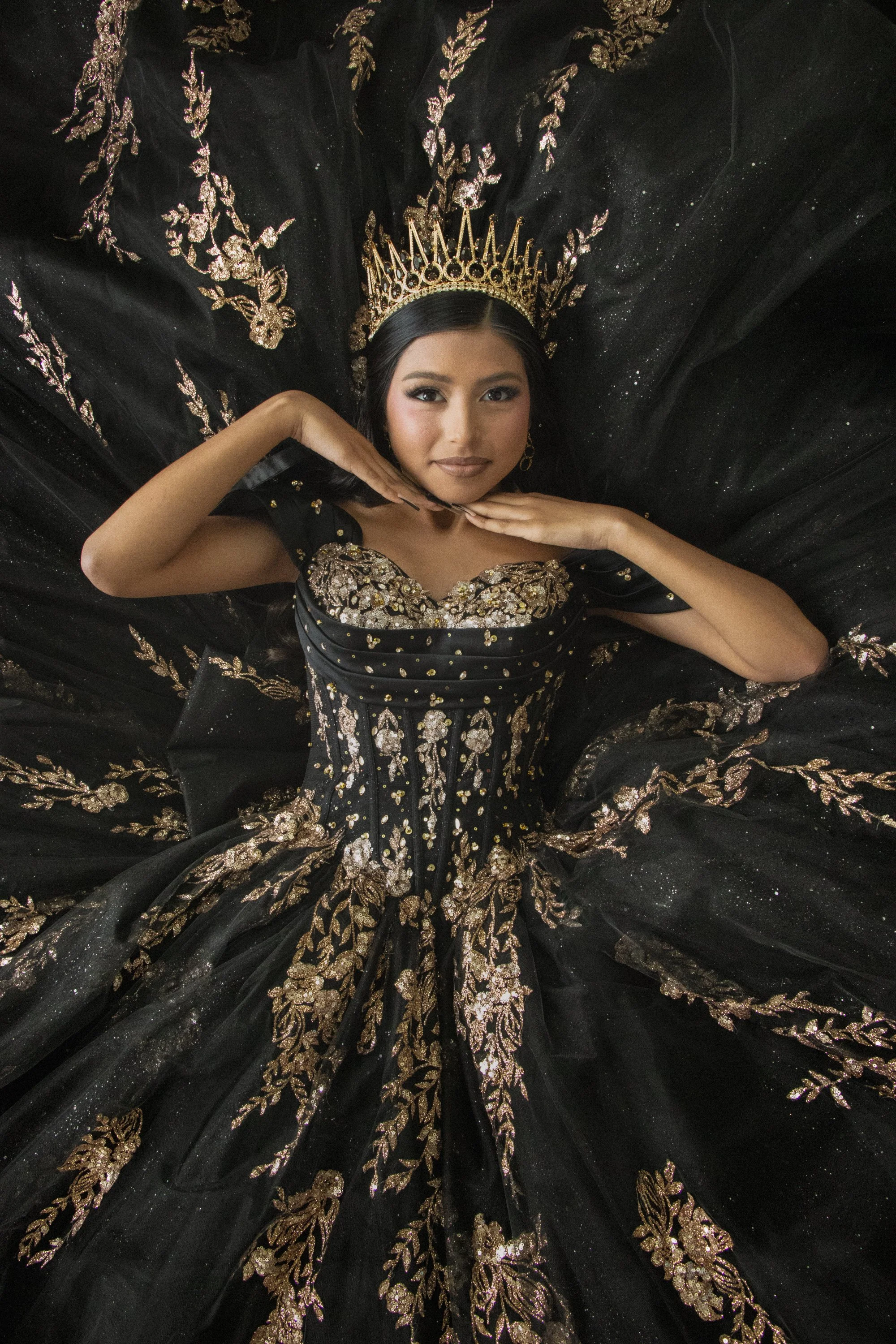 A woman wearing a black dress with intricate gold embroidery and a golden crown, lying down with her head supported by her hands on a matching black and gold embroidered fabric.