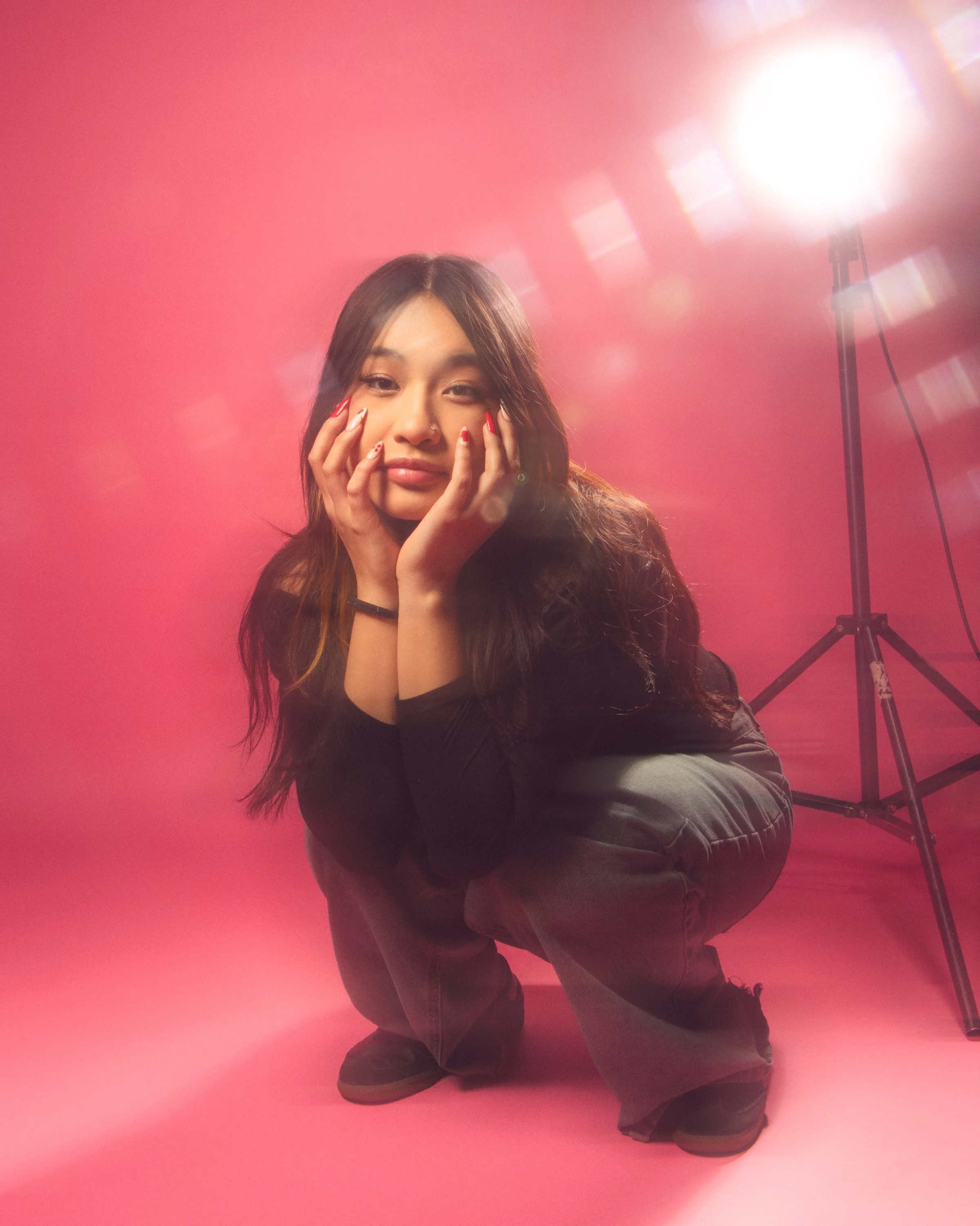A woman with long dark hair crouching on a pink floor in front of a pink background, with a bright studio light behind her to the right.
