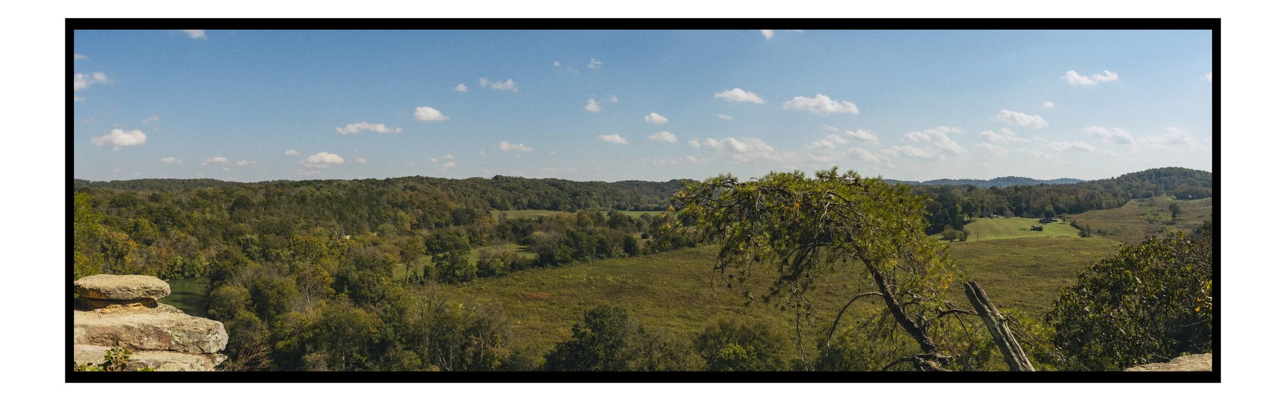 Scenic view of rolling hills with green trees and fields under a blue sky with scattered clouds.
