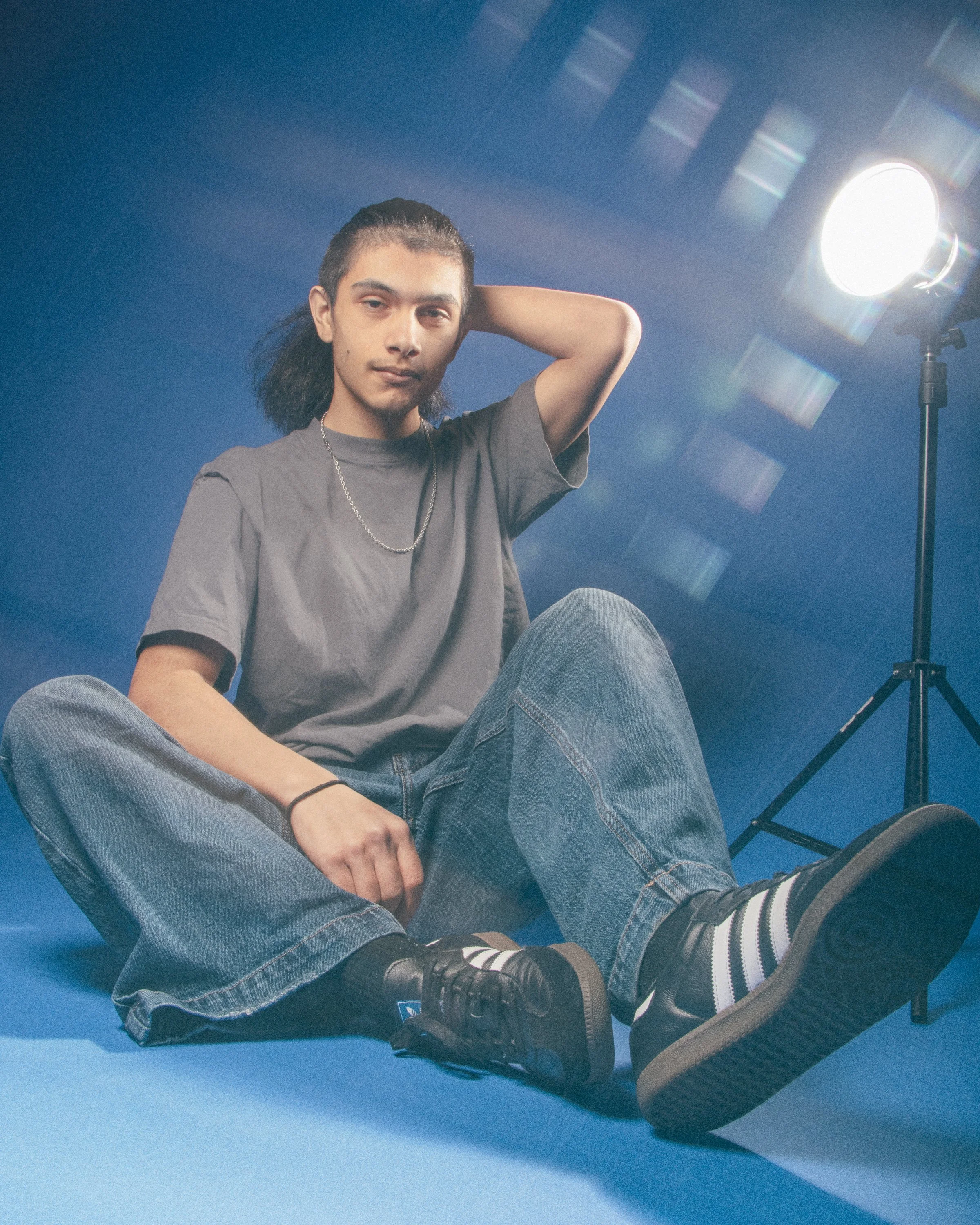 A young person sitting on the floor with legs crossed, wearing a gray t-shirt, jeans, black shoes, and striped socks, with one hand behind their head, in front of a blue background and studio lighting.