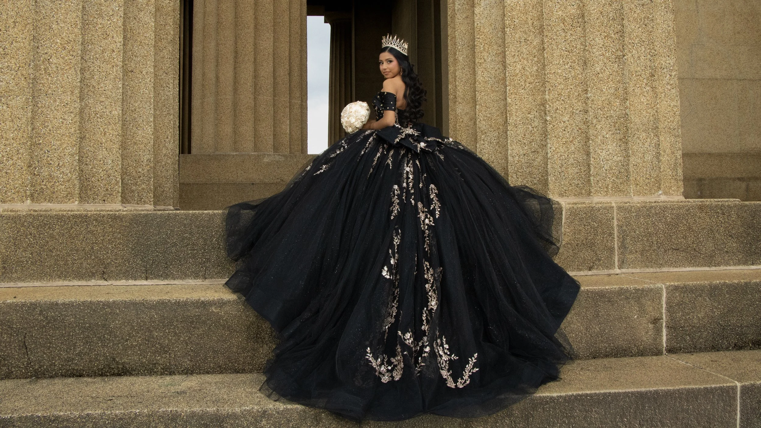 A woman in an elaborate black gown with white embroidery, wearing a silver tiara, holds a bouquet and stands on stone steps in front of a stone building, looking over her shoulder.