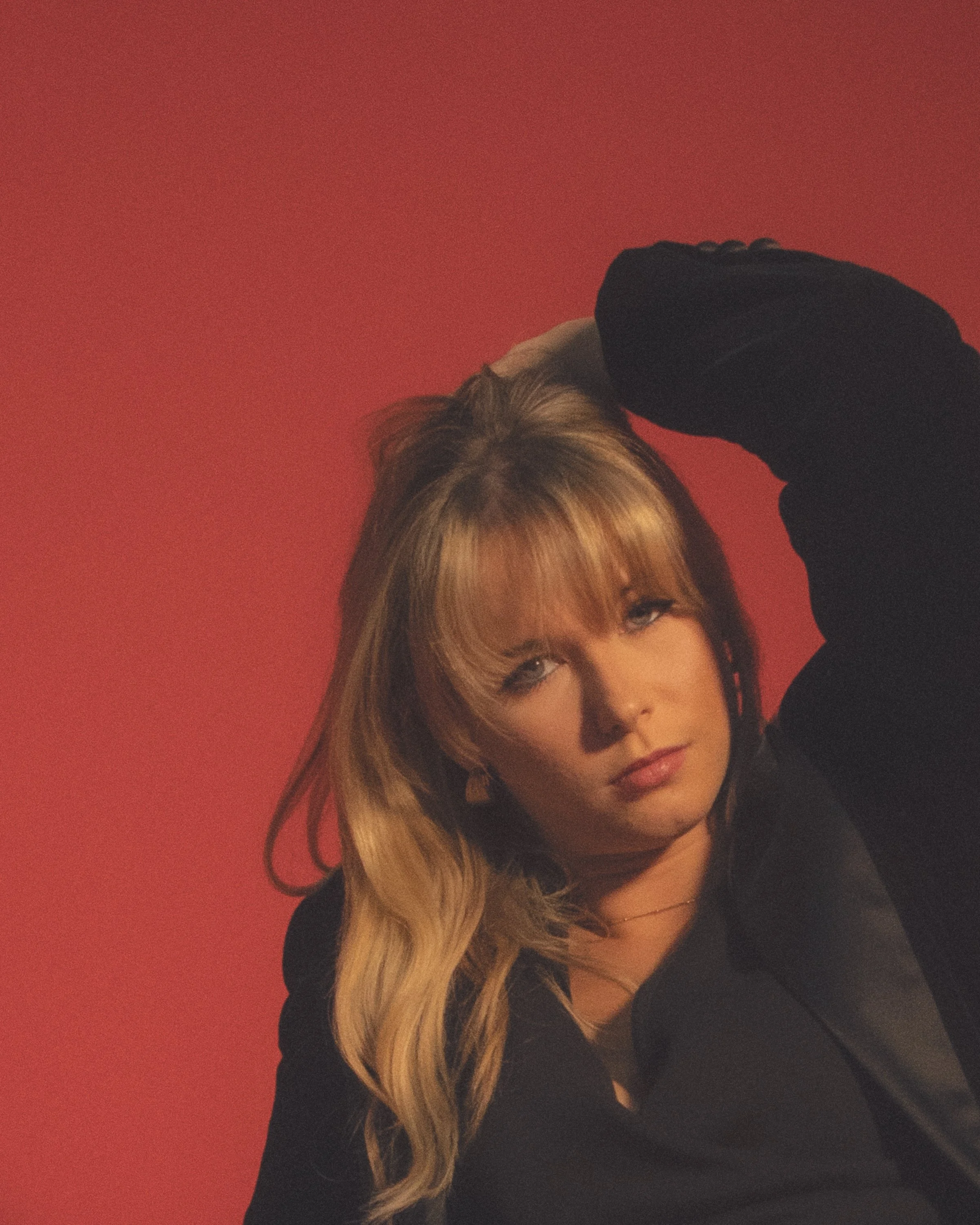 A woman with long blonde hair is posing with her hand on her head, looking at the camera, against a red background.