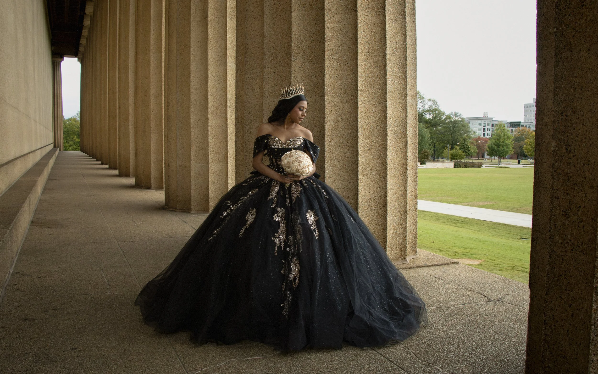 Young woman in a black and gold ball gown holding a bouquet of roses, wearing a crown, standing between stone columns in a park.