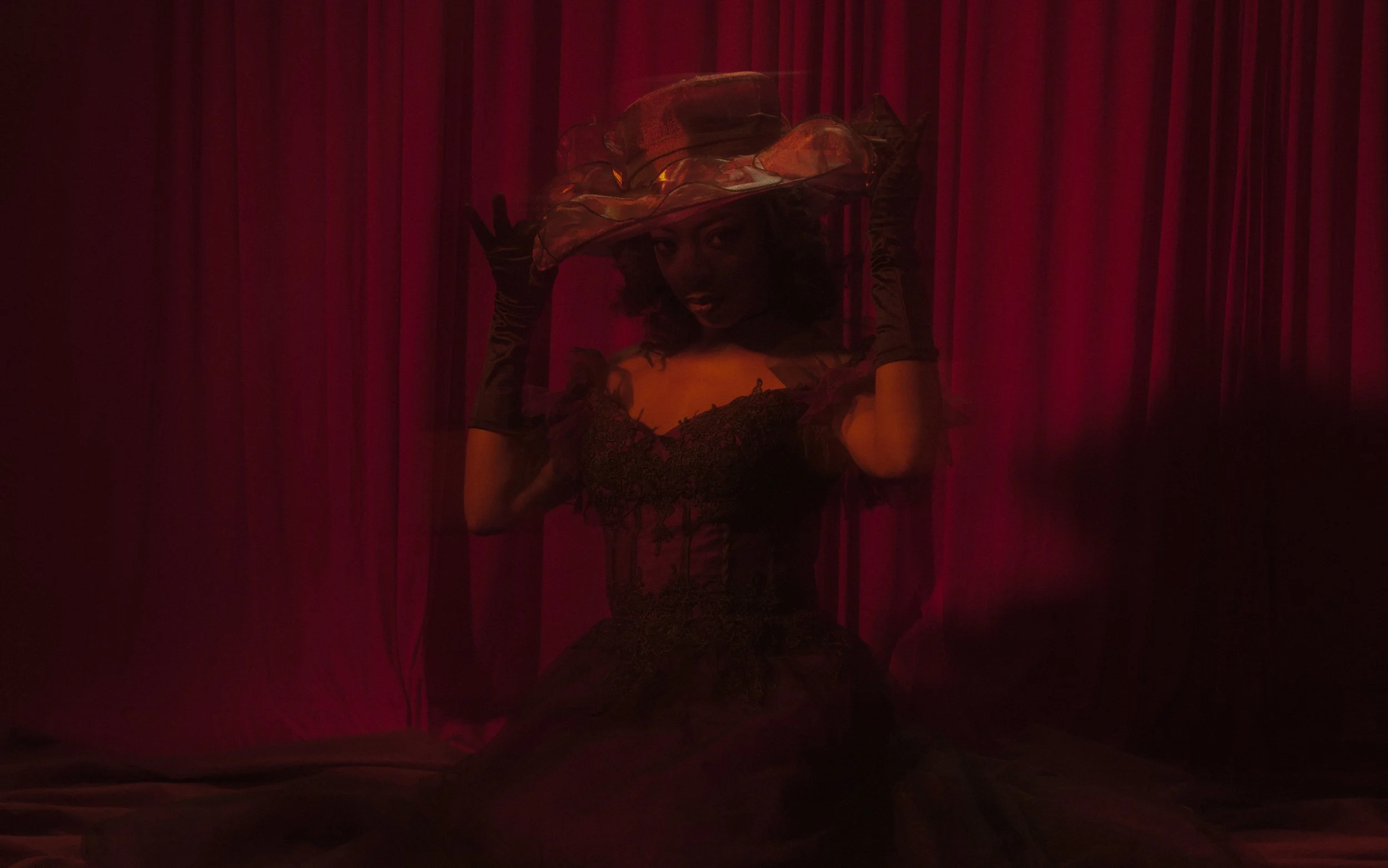 A woman wearing a large, colorful hat with pink, orange, and yellow hues, posing behind a semi-translucent pink curtain.