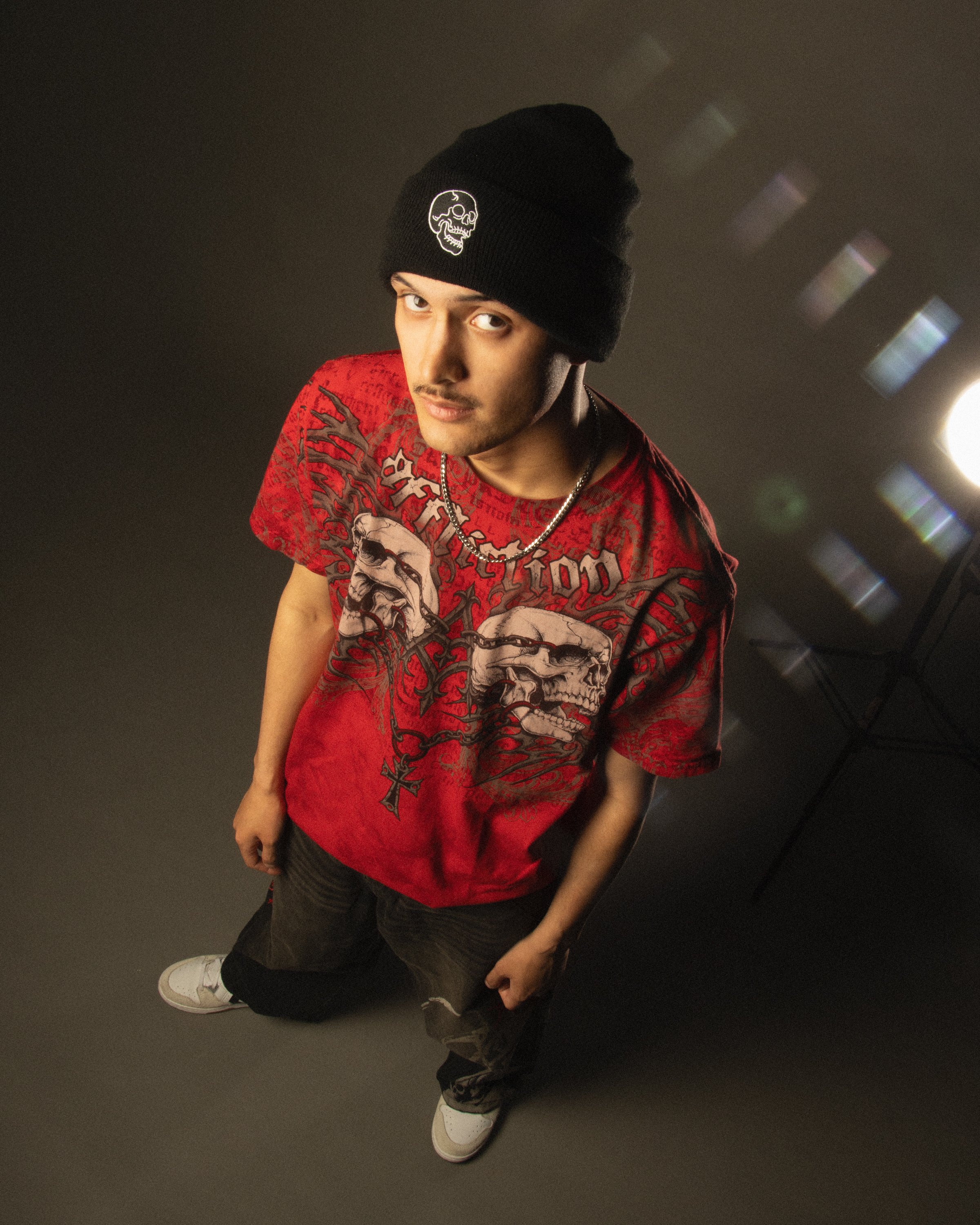 A young man wearing a black beanie with a skull design, a red graphic T-shirt, black pants, and white sneakers with gold accents, standing against a dark background.