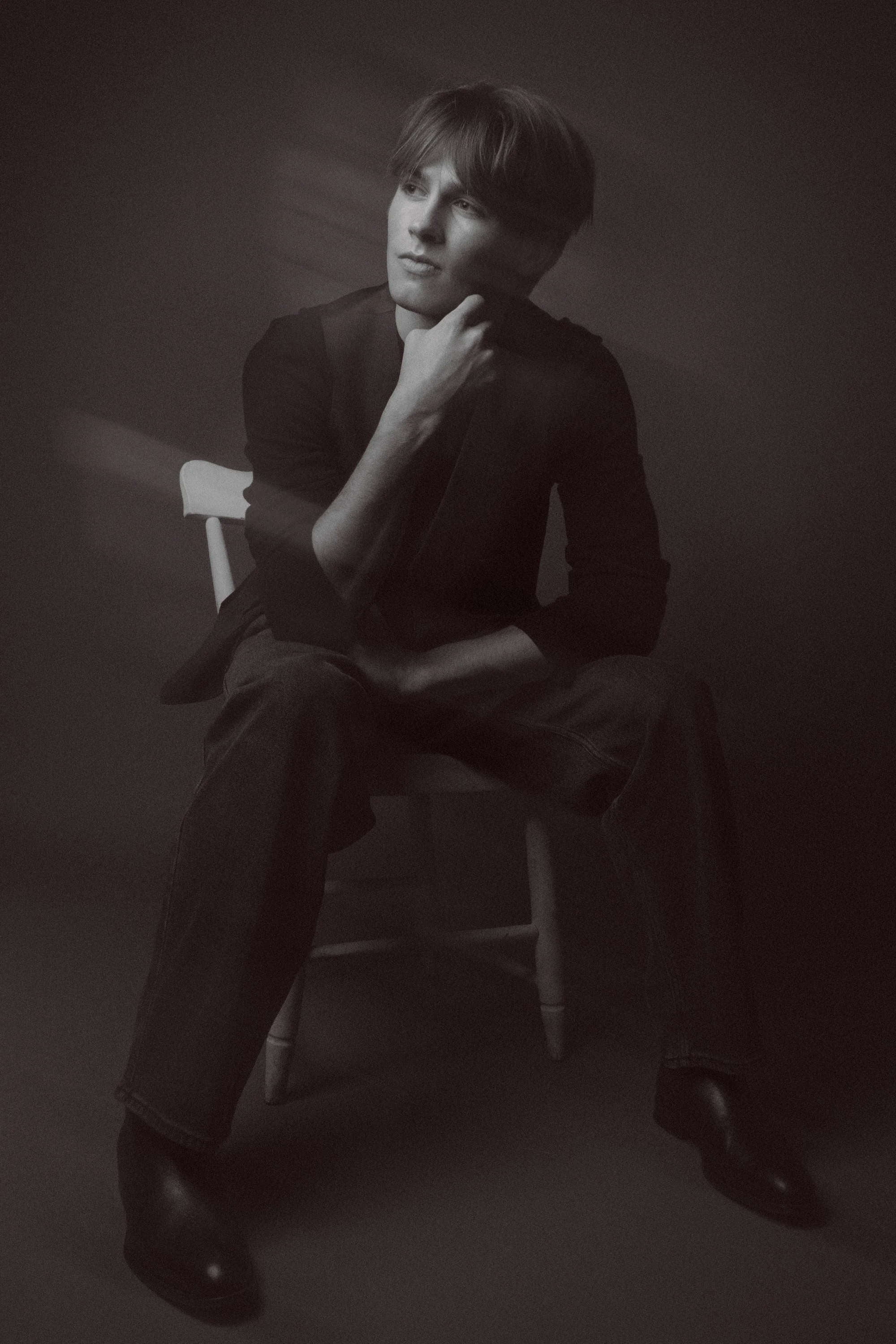 A young man with dark hair sitting on a white chair in a black shirt and pants, with a dark background.