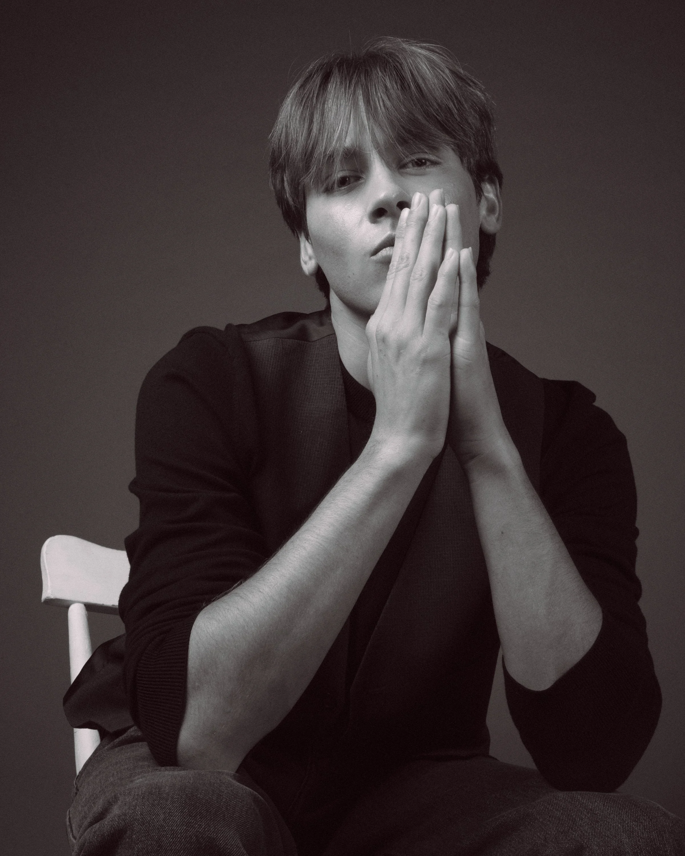 A black-and-white portrait of a young man with tousled hair, sitting on a white chair, resting his face on his hands, against a dark background.